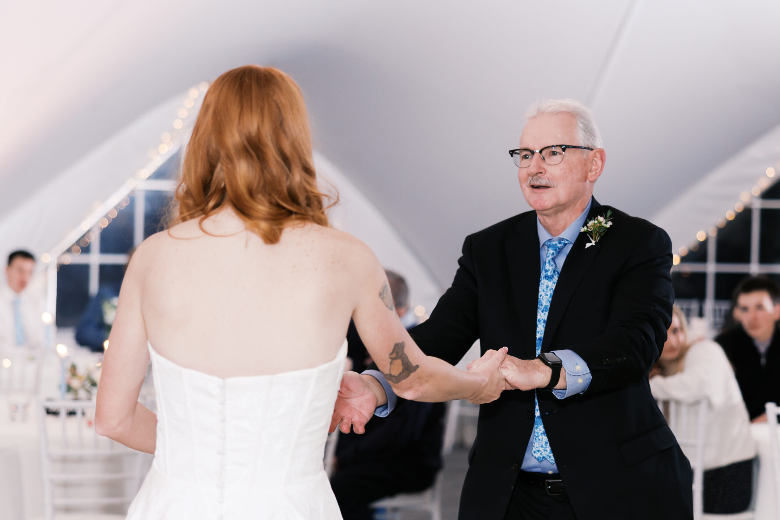 Bride and older man dancing at wedding reception with seated guests and soft lighting.