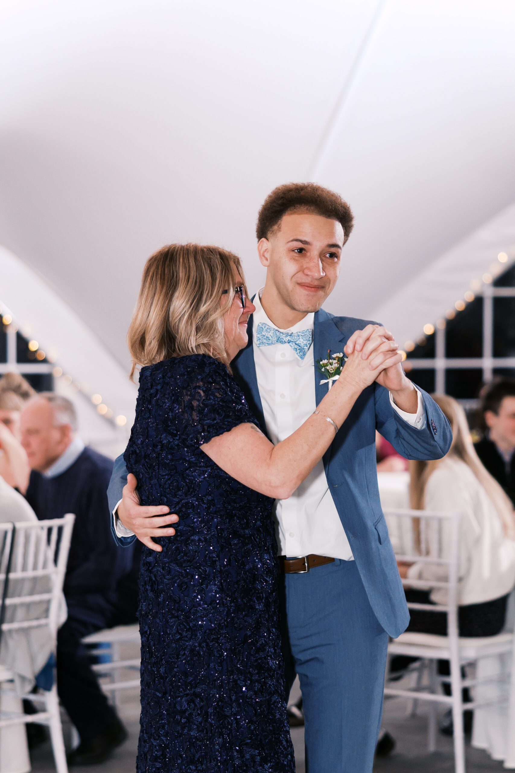 Groom dances with older woman in navy dress on reception dance floor surrounded by seated guests.