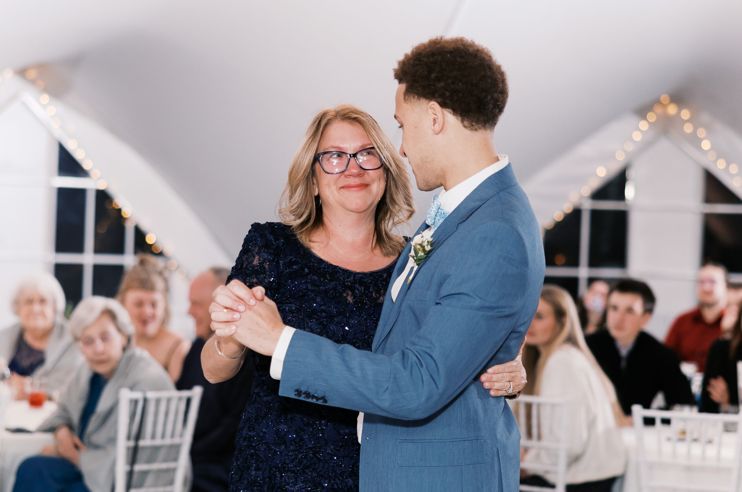 Groom and older woman share emotional dance moment at wedding reception with guests in background.