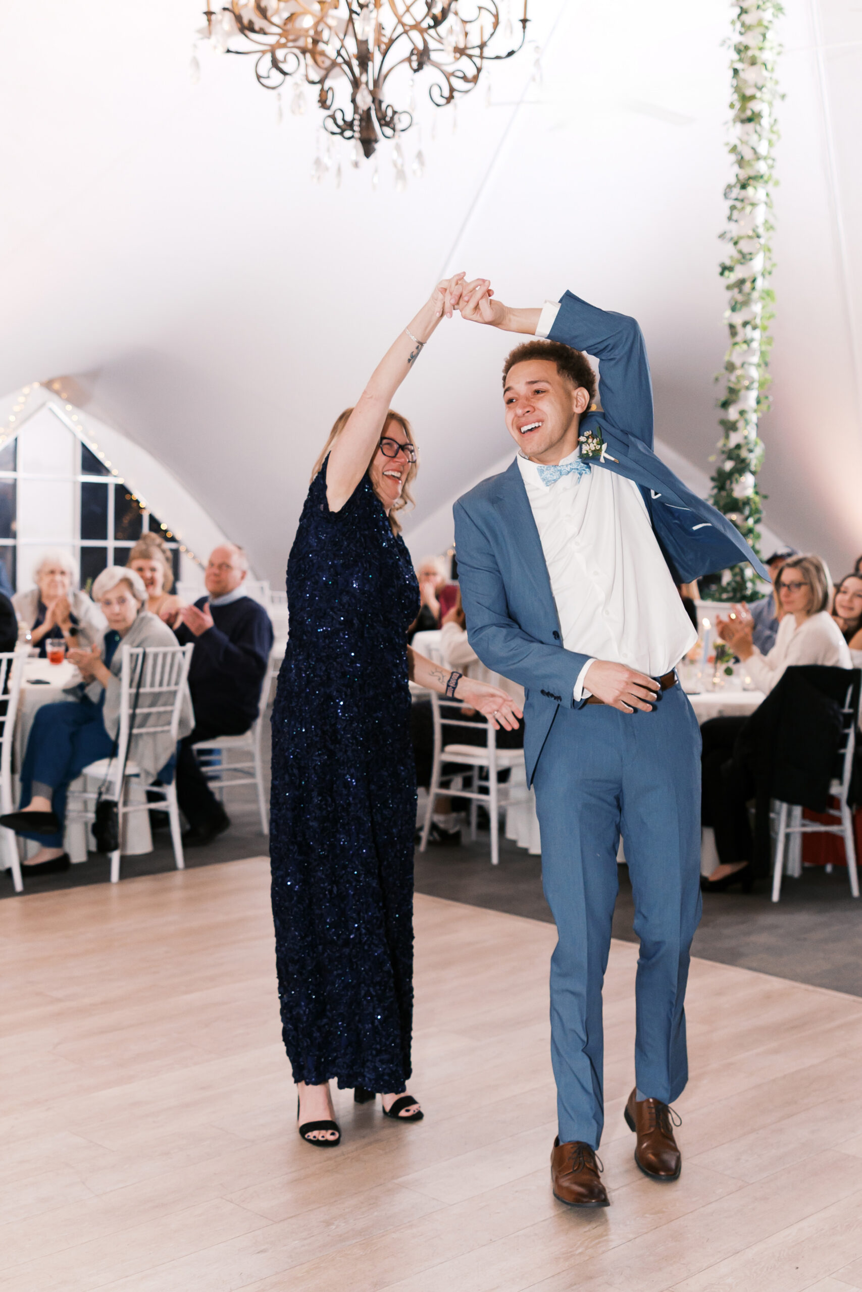 Groom spins older woman on dance floor under chandelier at wedding reception with applauding guests.