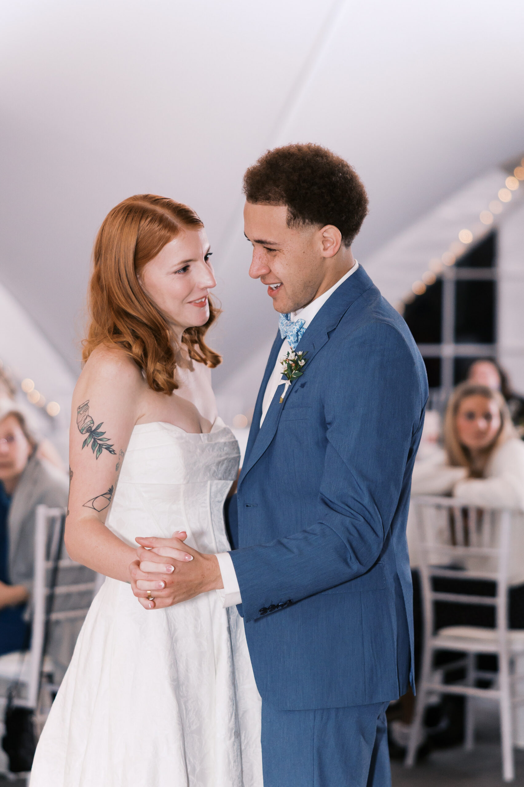 Bride and groom share first dance on reception dance floor with guests seated around them.