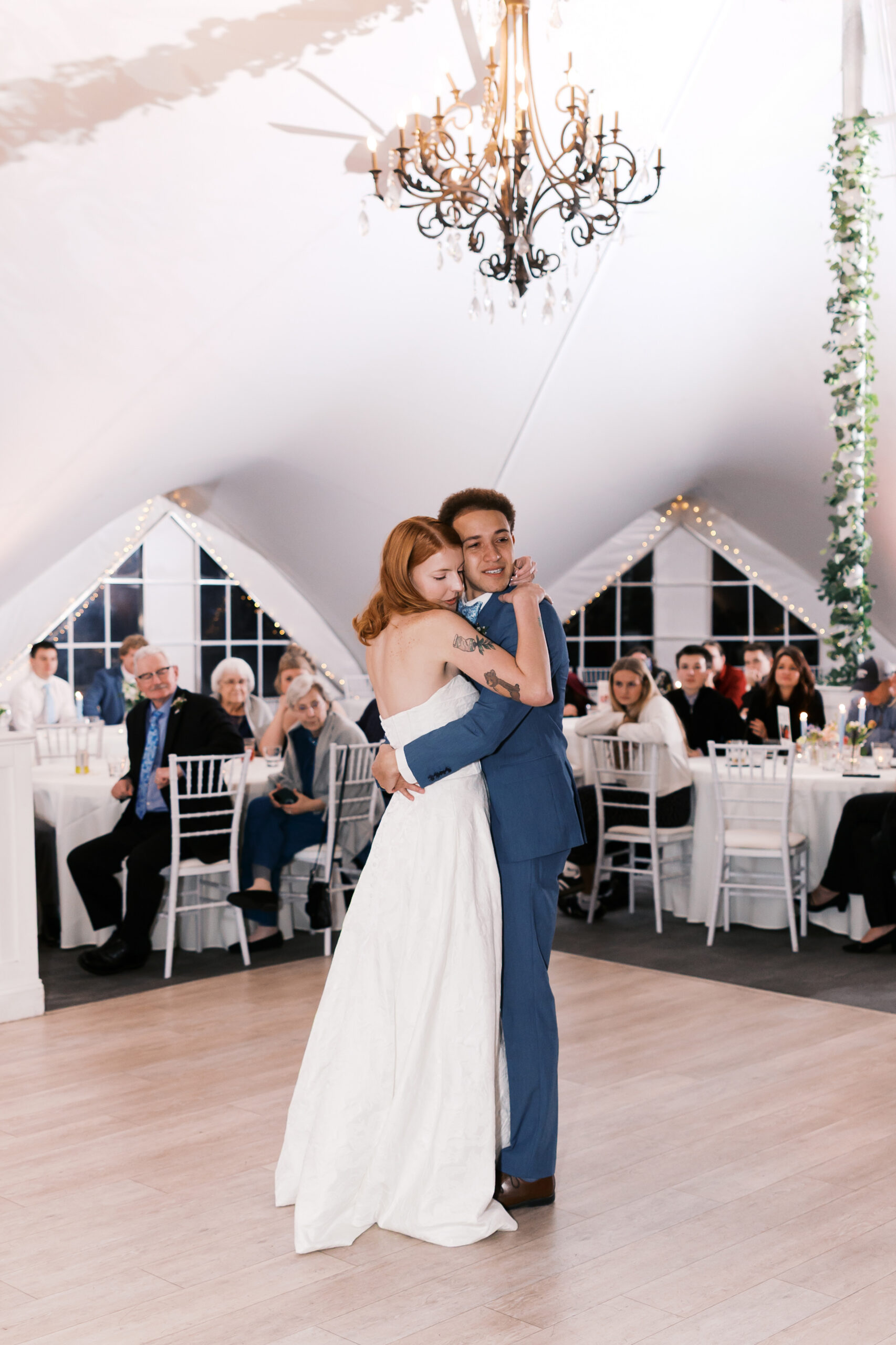 Bride and groom hug during first dance on reception dance floor with guests seated around them.