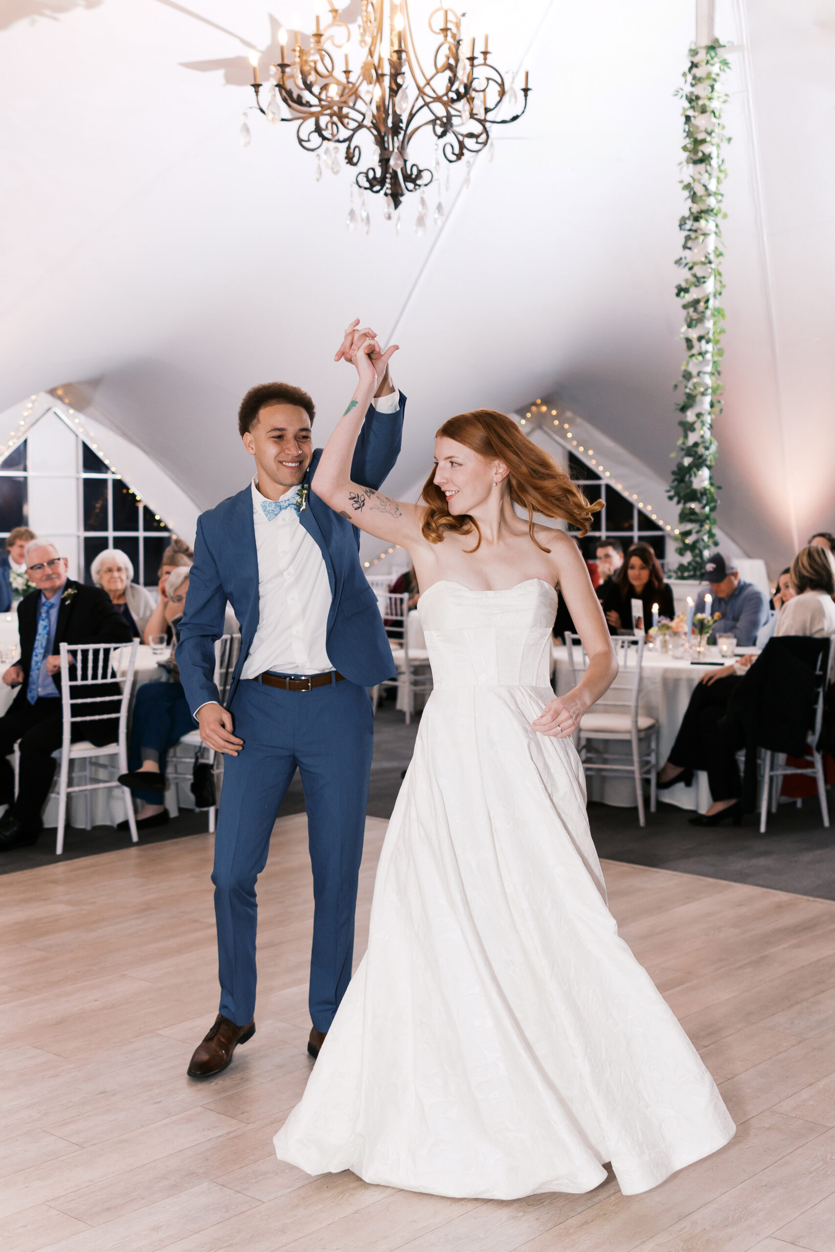 Bride and groom spin together on dance floor under chandelier and string lights at wedding reception.