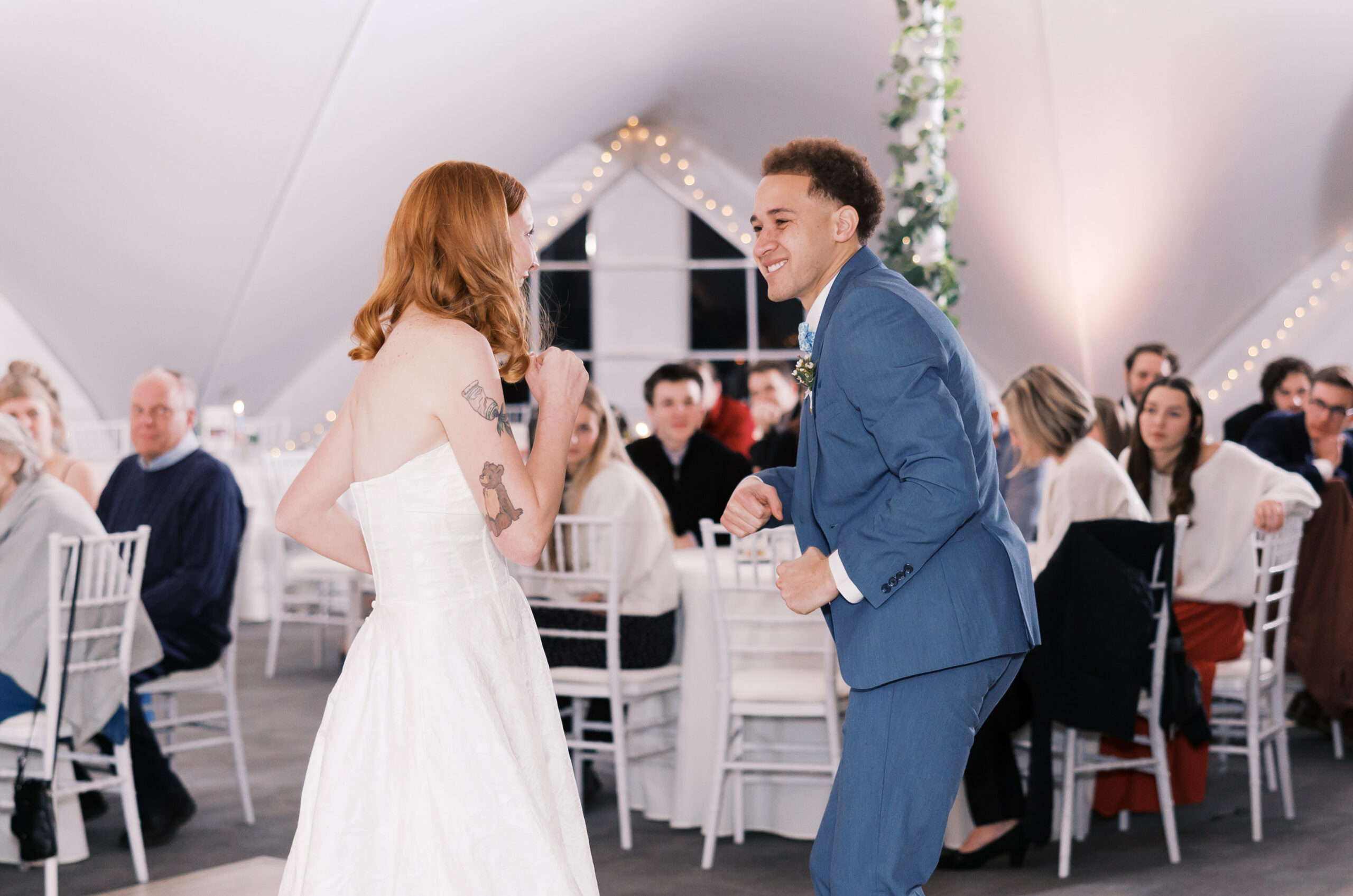 Bride and groom laugh and dance together on reception dance floor with guests watching nearby.