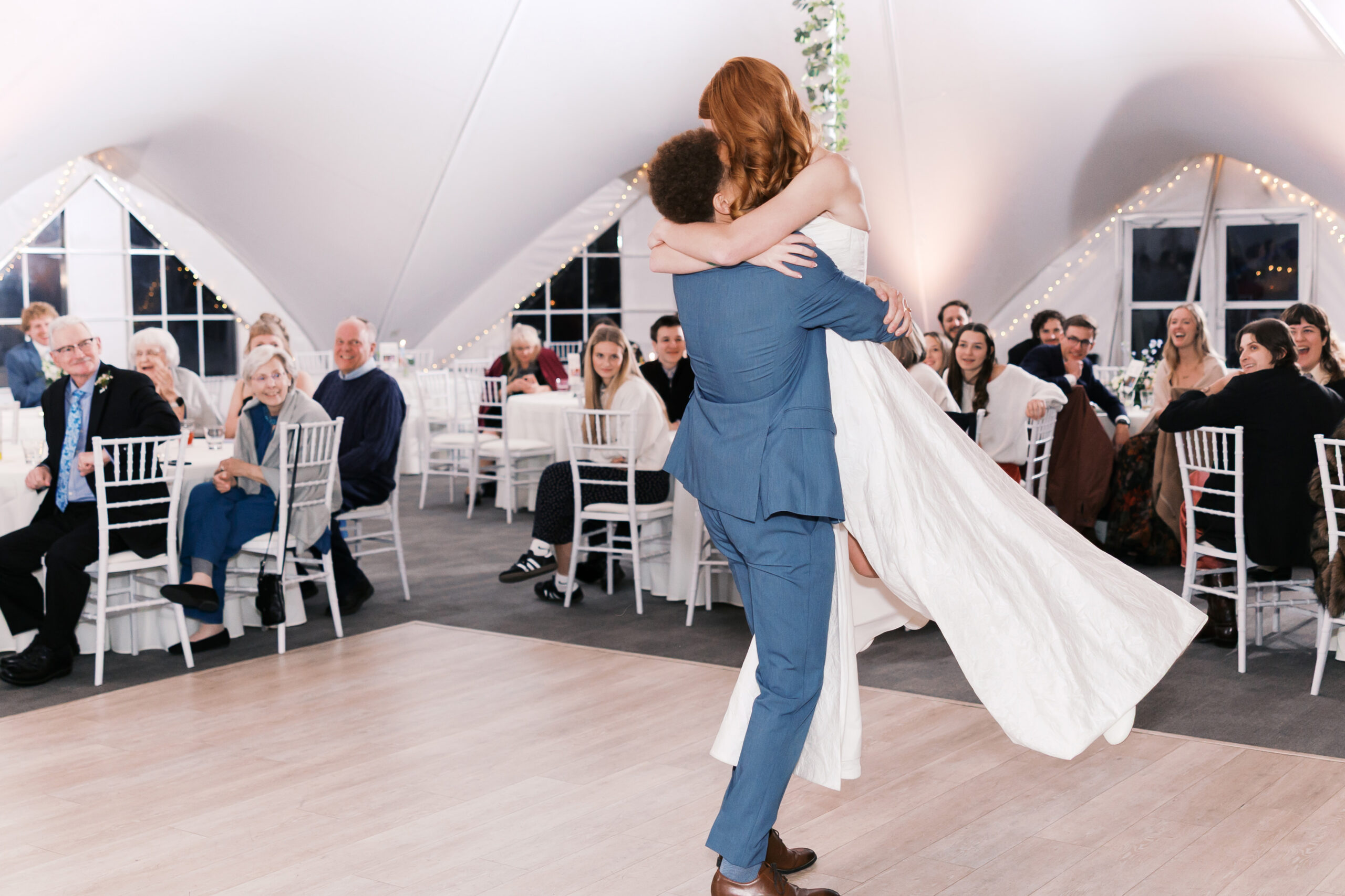 Groom lifts bride during first dance on reception dance floor surrounded by smiling guests.