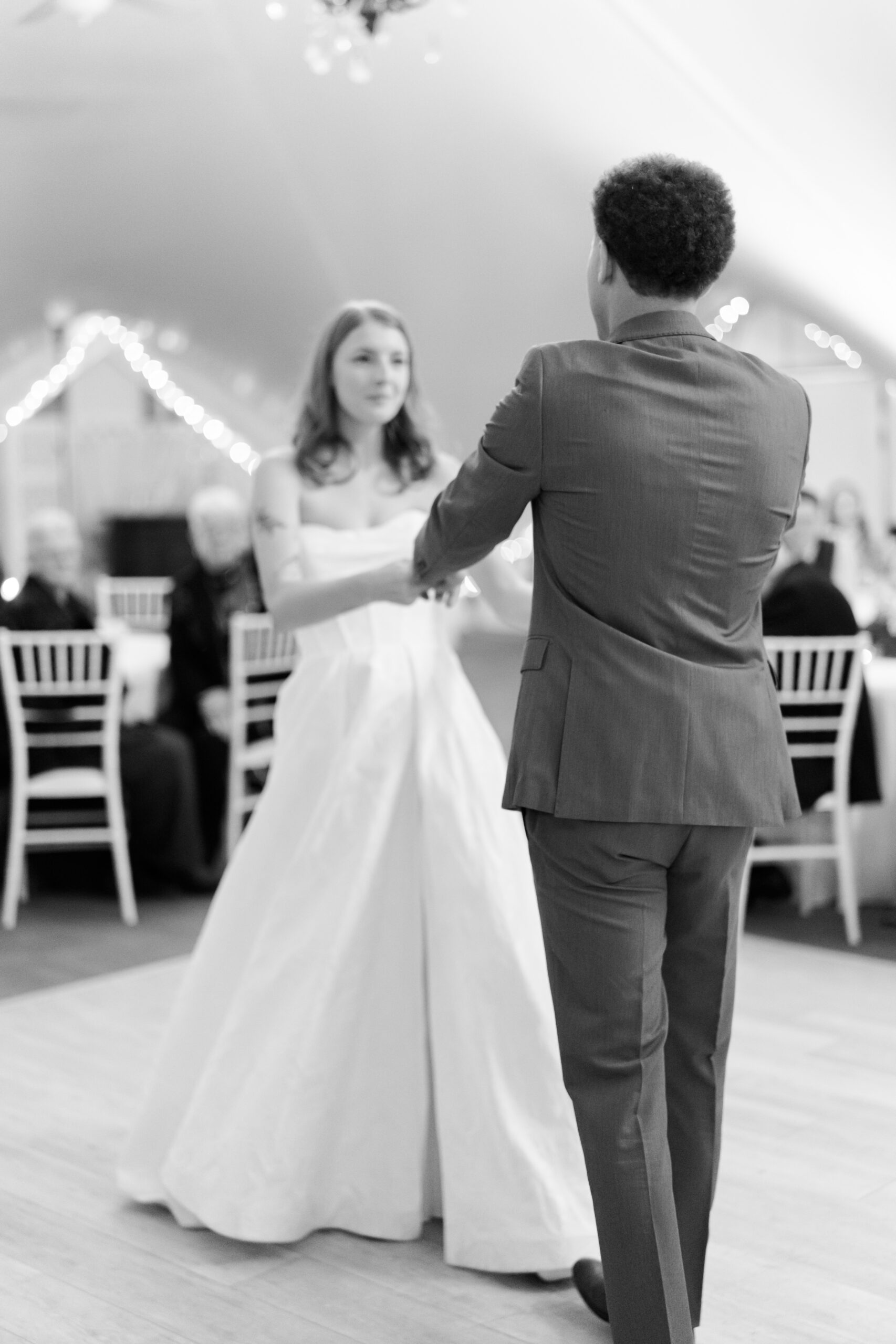 Bride and groom dance together on reception floor in black and white photo with guests seated around.
