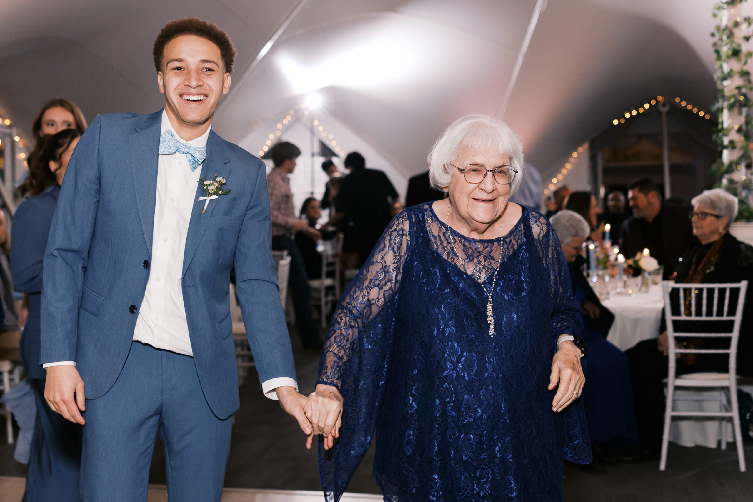 Groom walks hand in hand with elderly woman in navy lace dress at wedding reception.