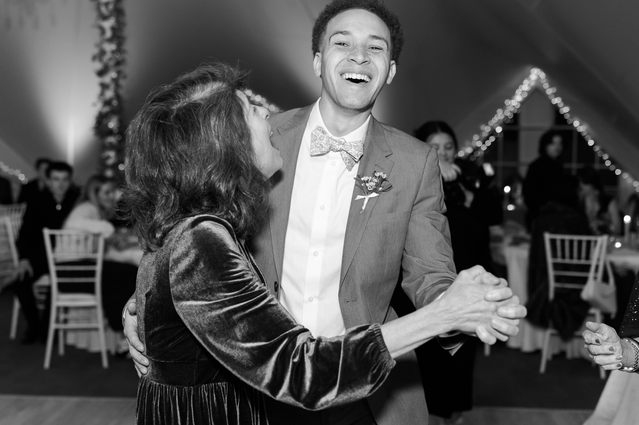 Groom dances with older woman on reception dance floor in black and white candid moment.