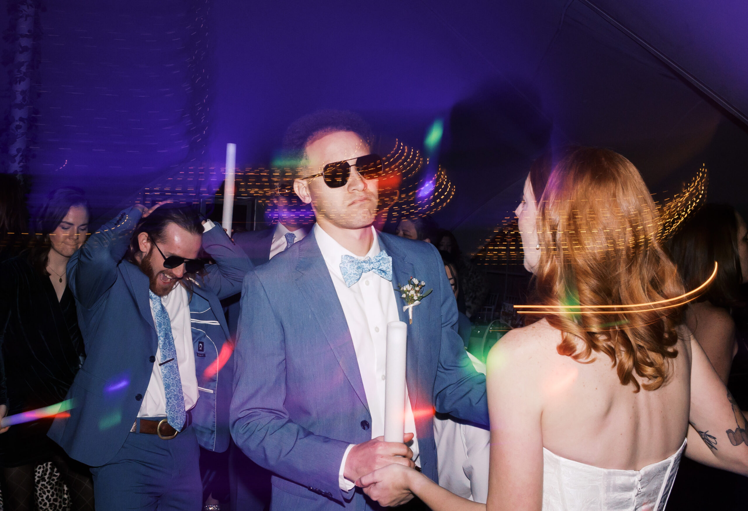 Guests dancing at wedding reception with glow sticks and colorful light trails, groom in blue suit and sunglasses facing bride