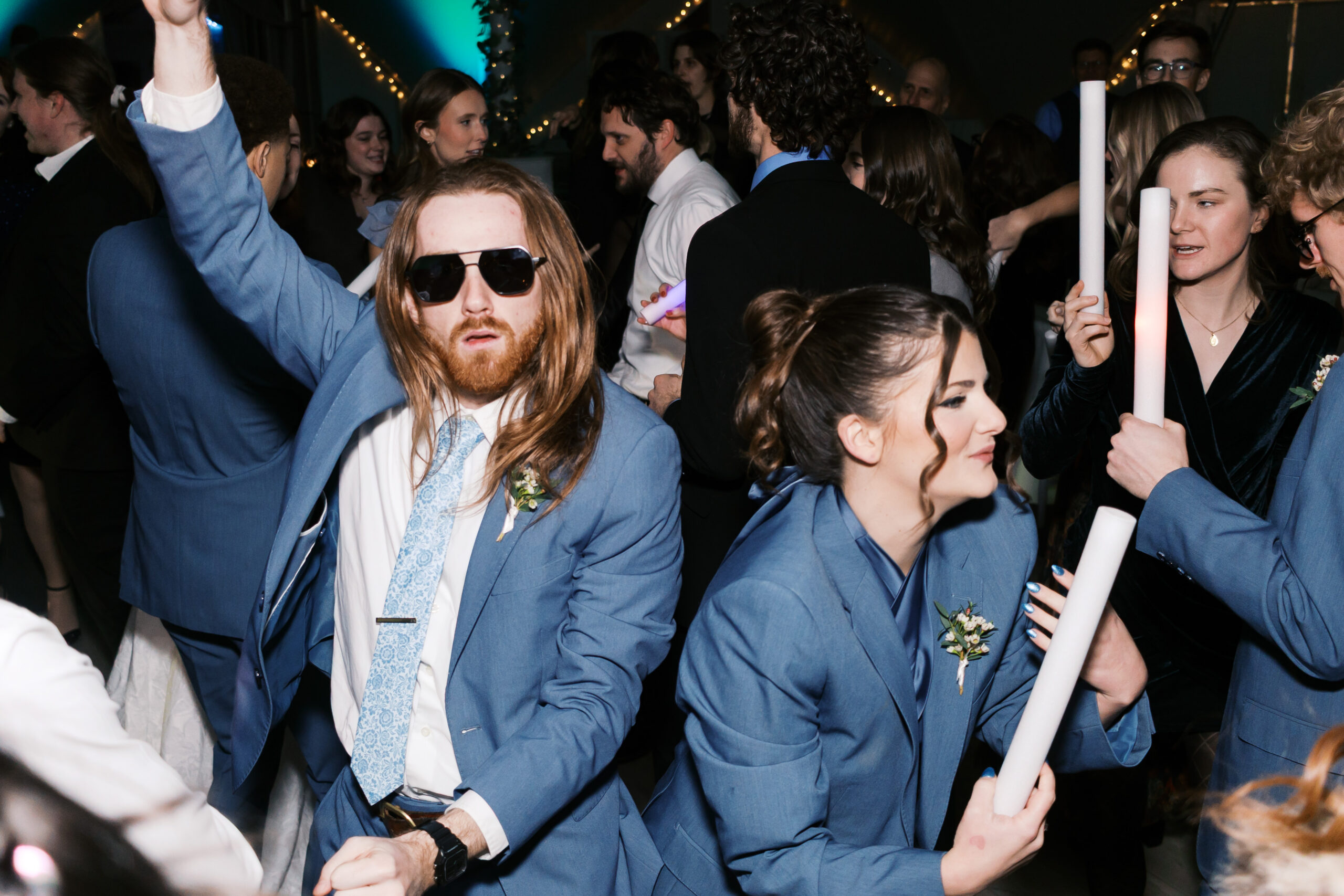 Groomsman in blue suit and sunglasses dancing energetically on crowded wedding dance floor with glow sticks