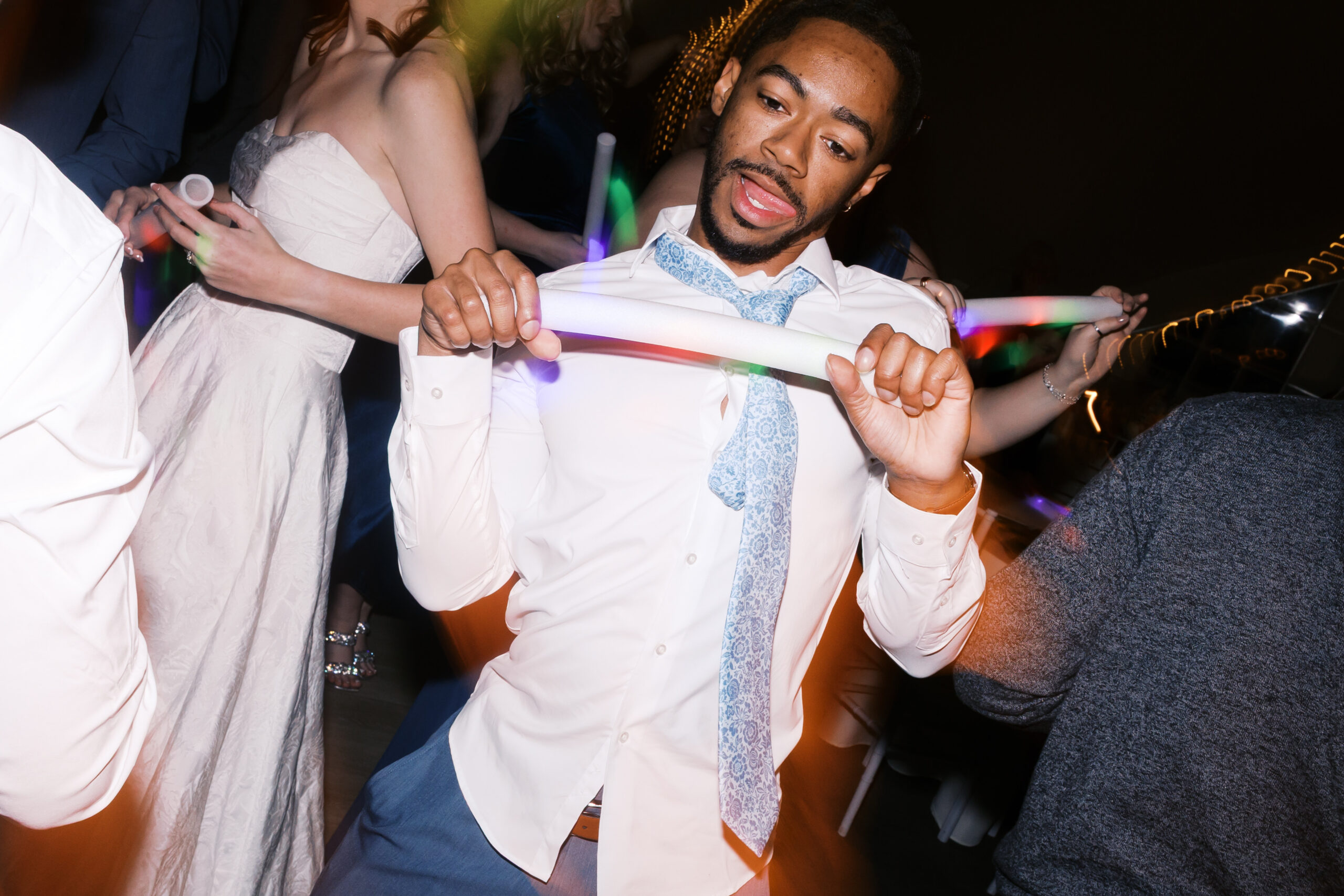 Wedding guest in white shirt and loosened tie dancing while holding glowing foam stick on reception dance floor
