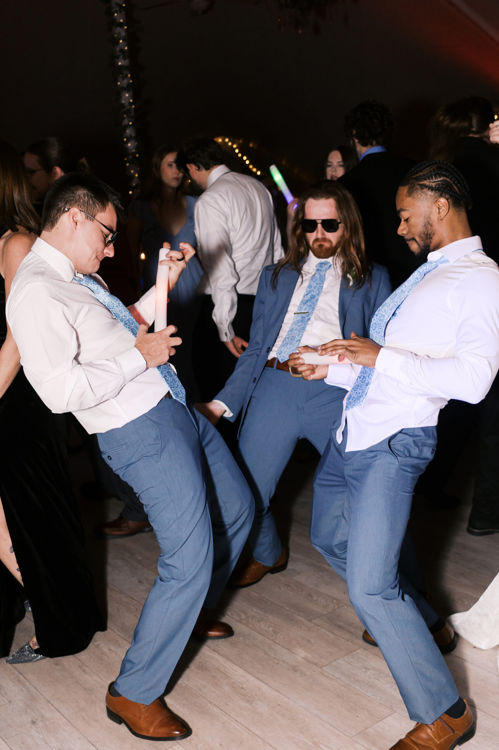 Three groomsmen in blue suits dancing together with glow sticks on wedding reception dance floor