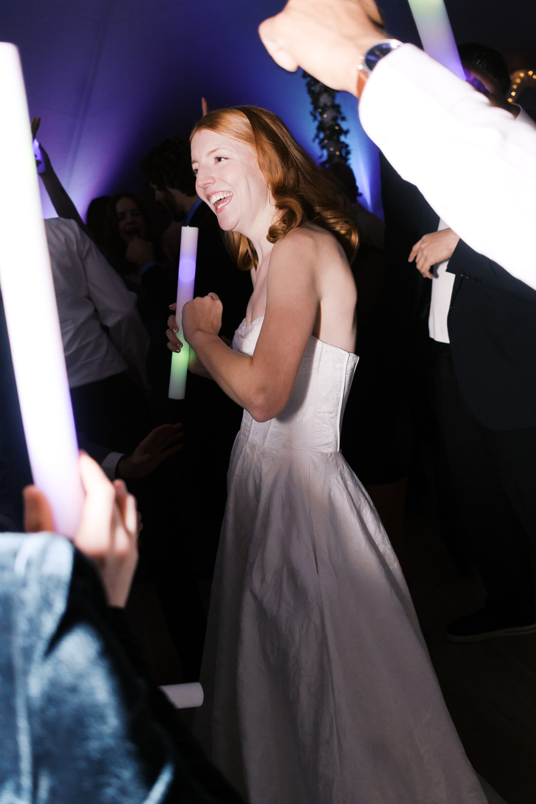 Bride in strapless gown smiling and dancing with glowing foam stick surrounded by guests
