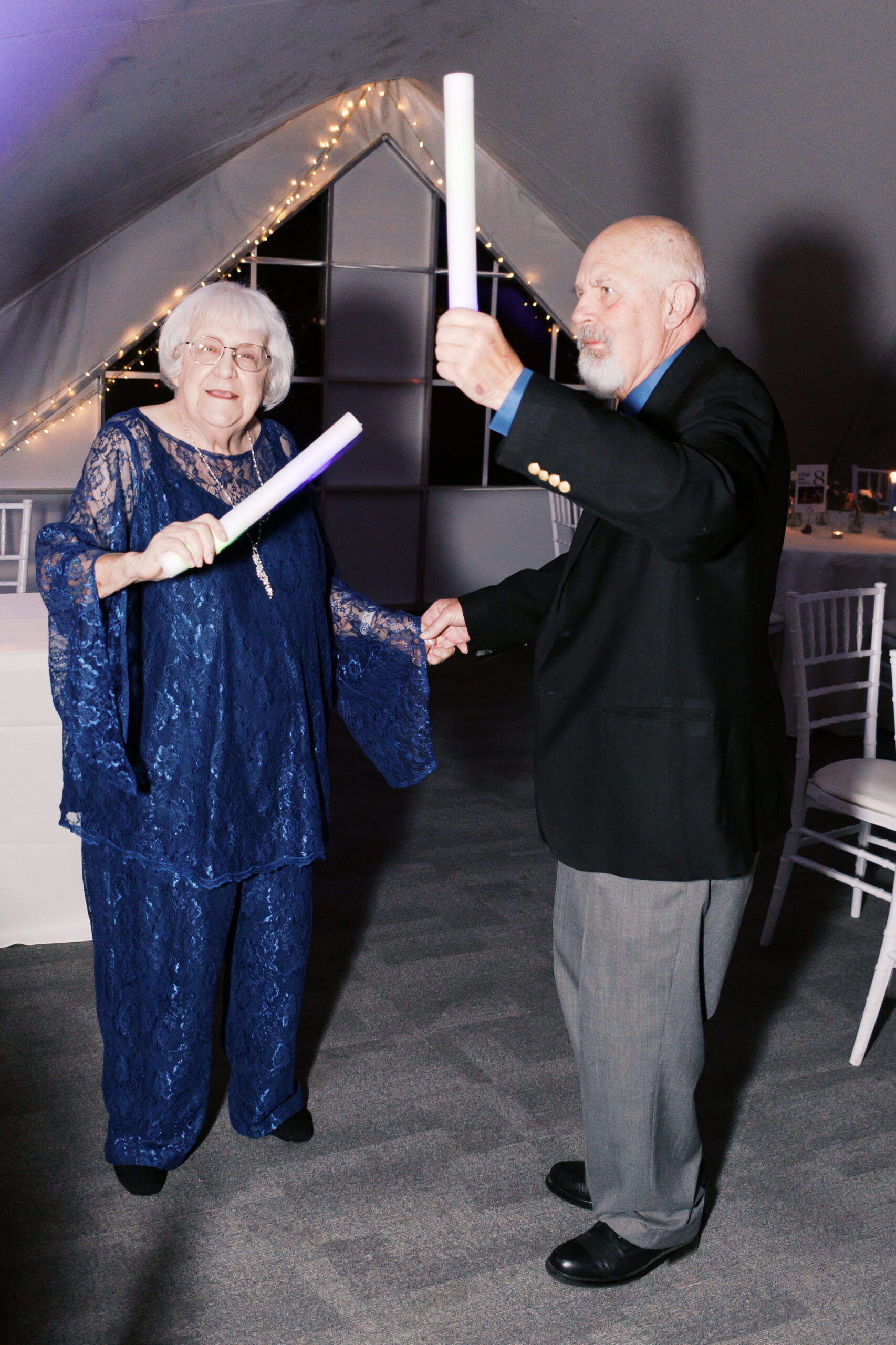 Elderly couple dancing together holding glow sticks inside softly lit wedding tent