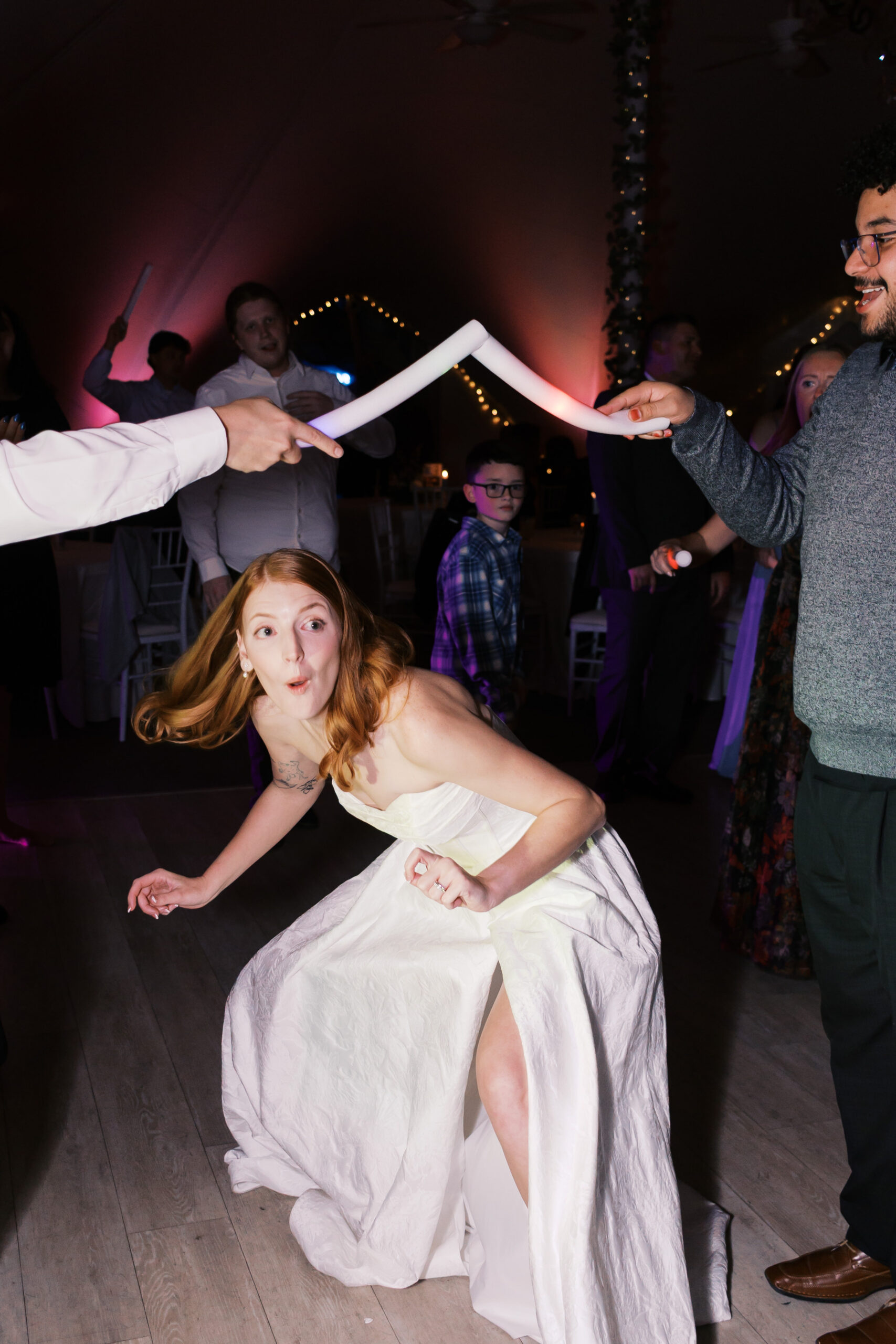 Bride crouching playfully on dance floor during reception game while guests watch and hold glow sticks
