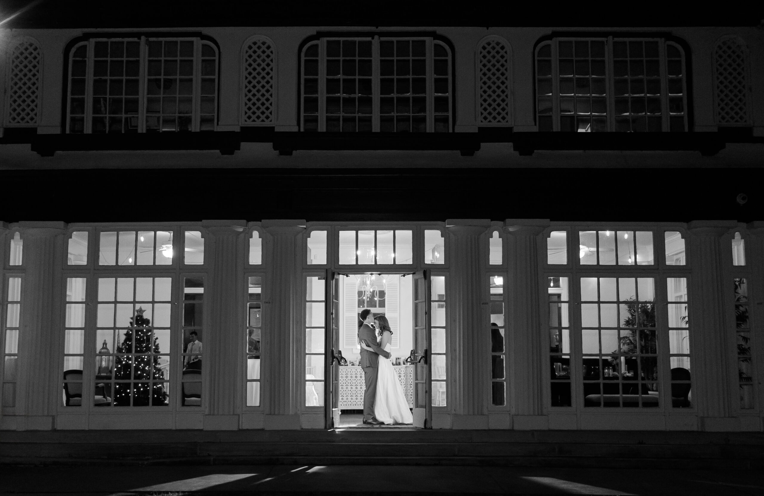 Couple kissing in doorway of lit reception venue at night, framed by large windows and interior lights