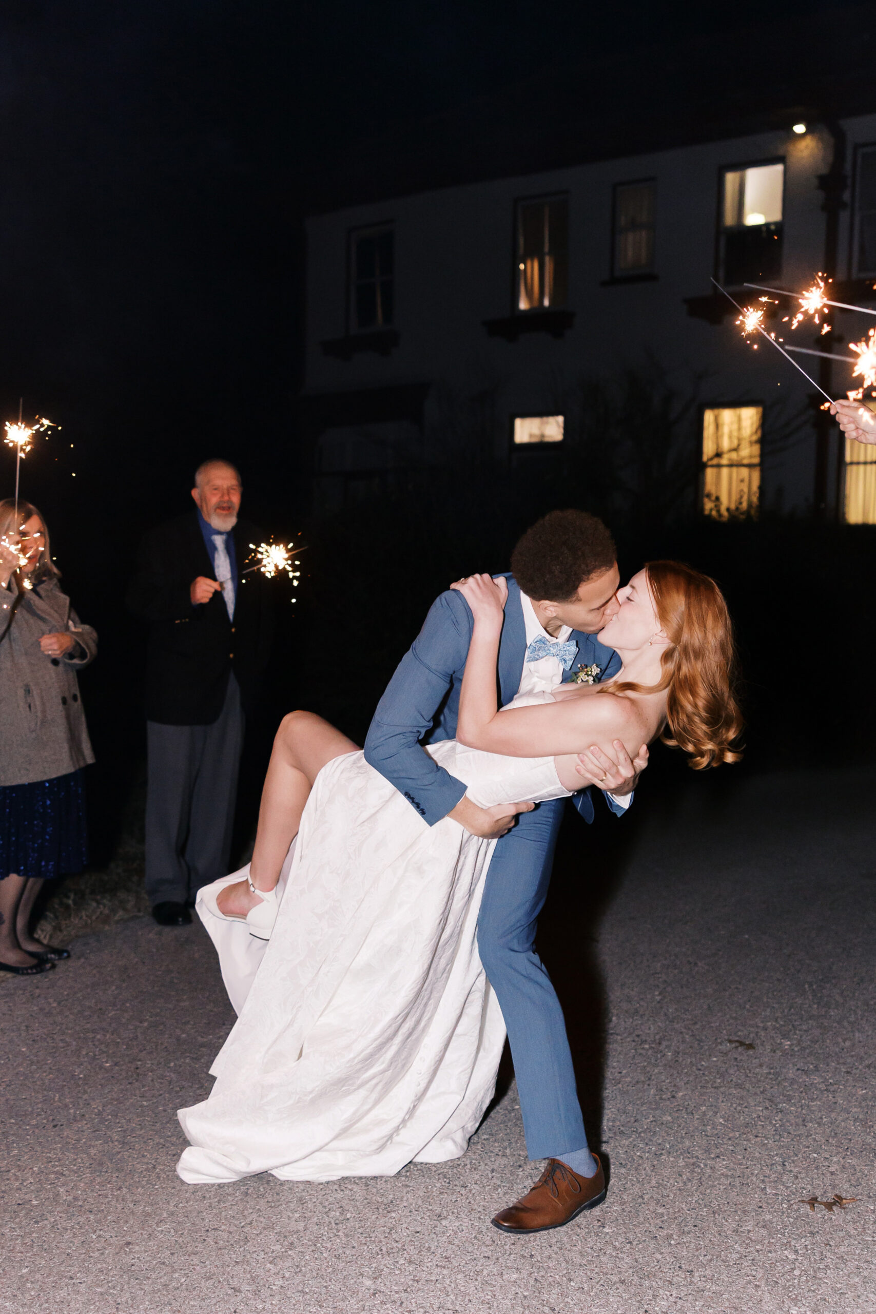 Groom dips bride for a kiss outside at night while guests hold sparklers during wedding send-off