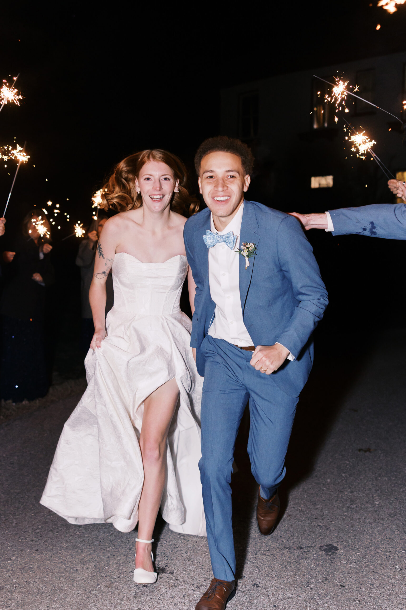 Bride and groom running together through sparkler exit at night, smiling as guests celebrate behind them