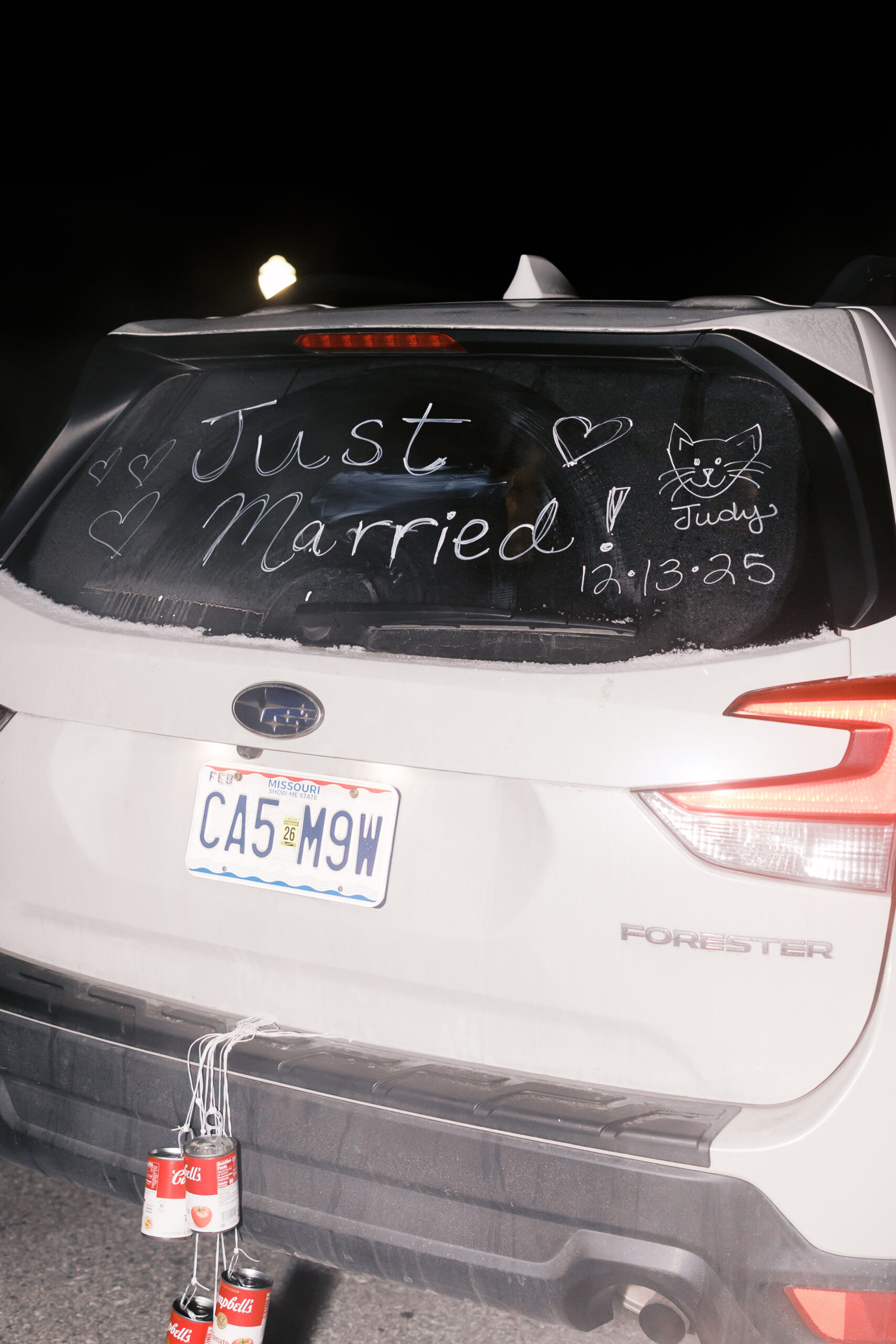 White getaway car with “Just Married” written on rear window and cans attached to bumper at night