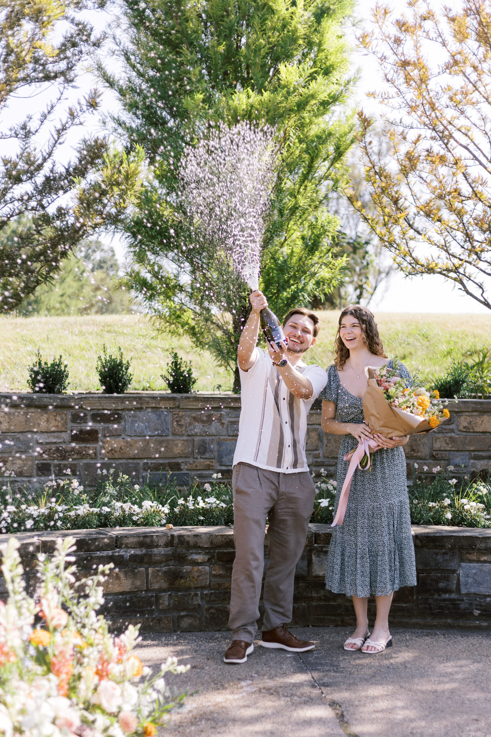 Man pops champagne while woman holds bouquet, celebrating proposal at Powell Gardens garden.