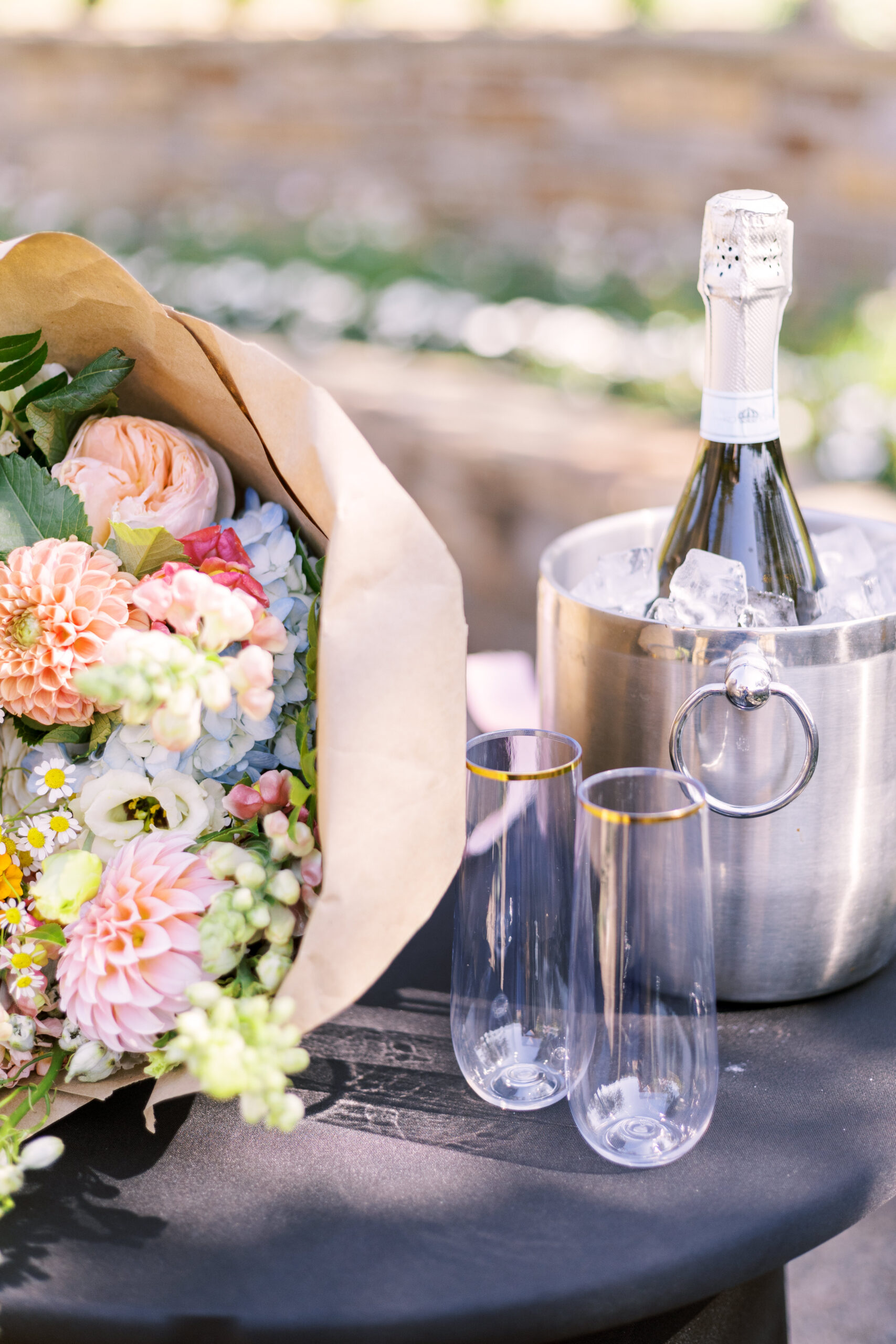 Bouquet and champagne bottle in ice bucket with two glasses on outdoor table at Powell Gardens.