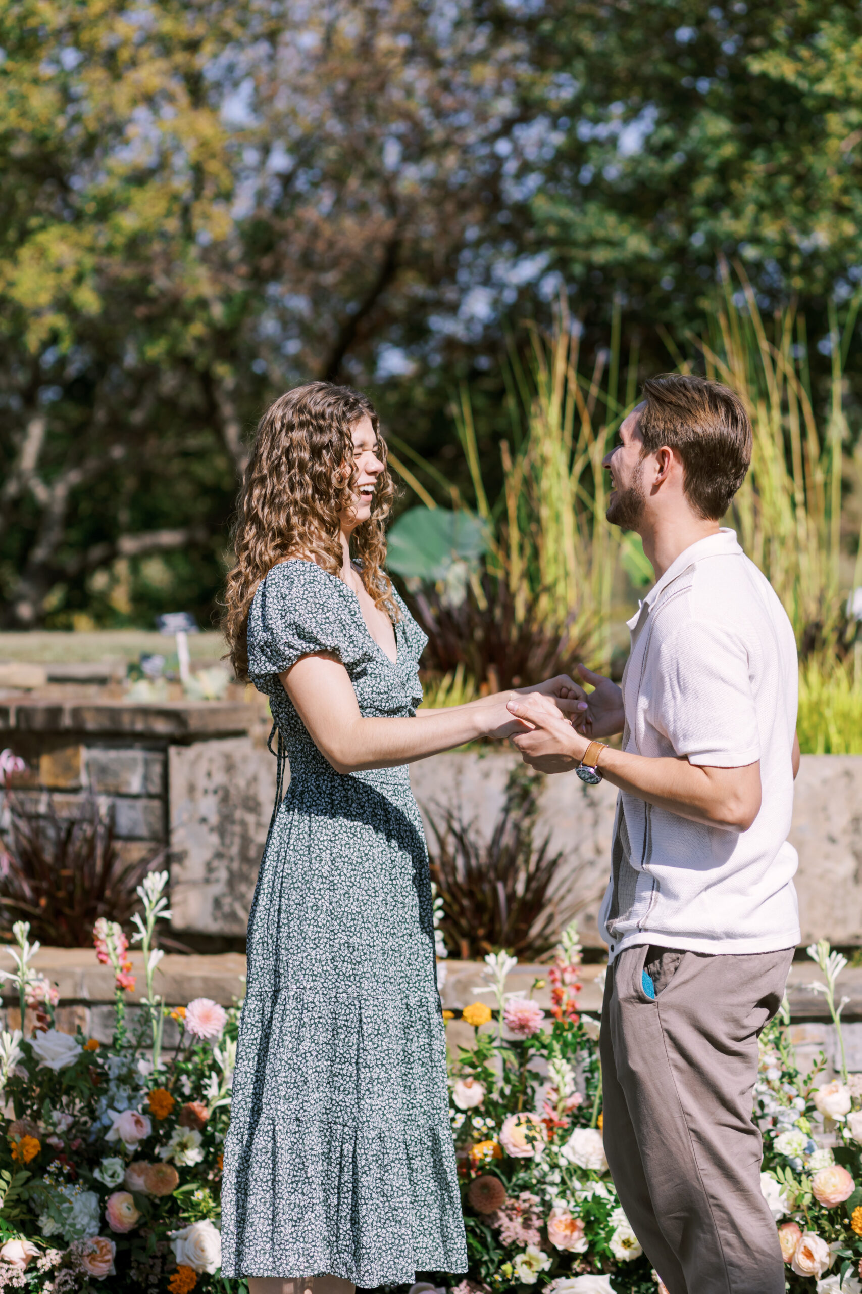 Couple holding hands and smiling at each other after proposal in garden setting.