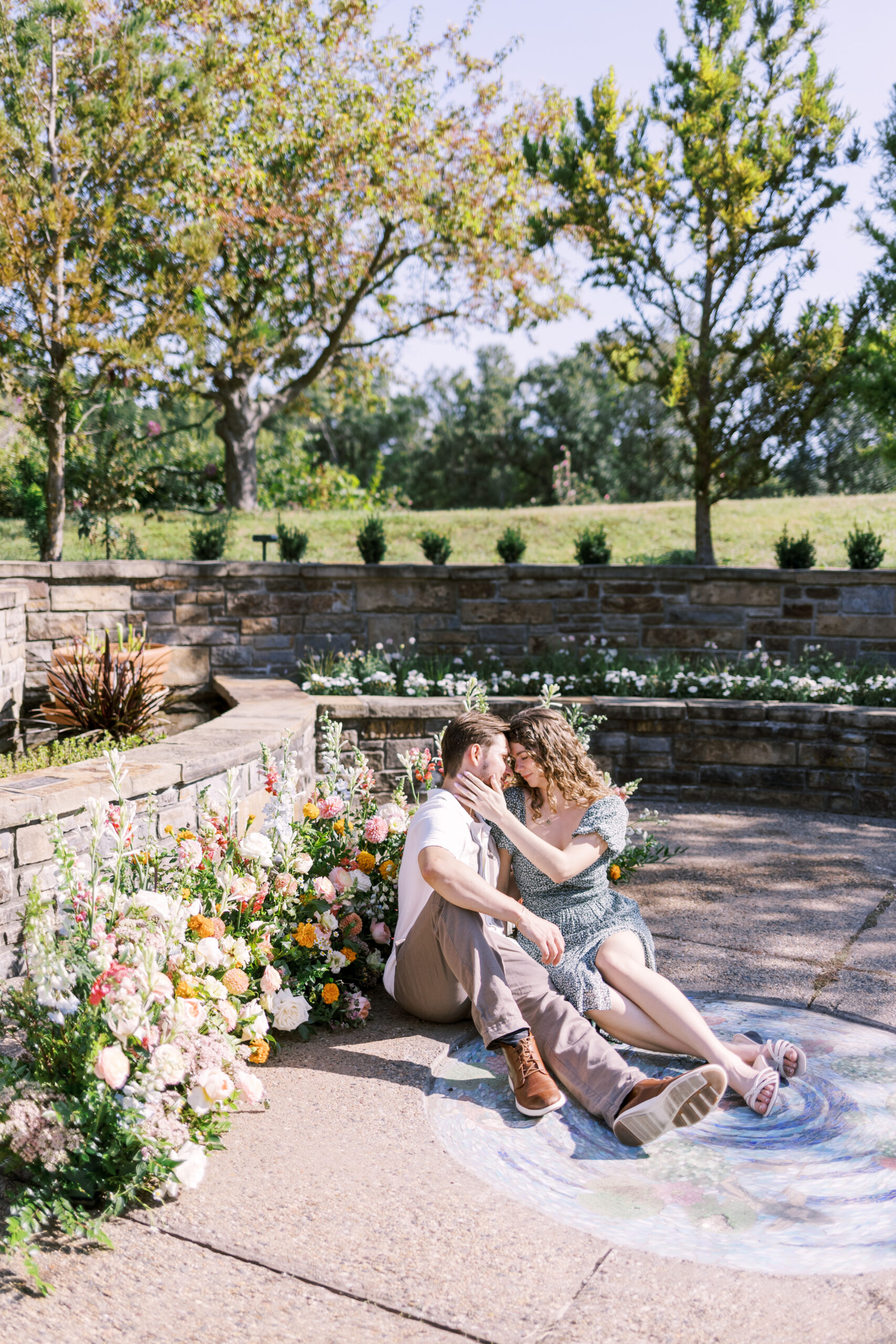 Newly engaged couple sitting together beside floral arrangements in sunny garden space.