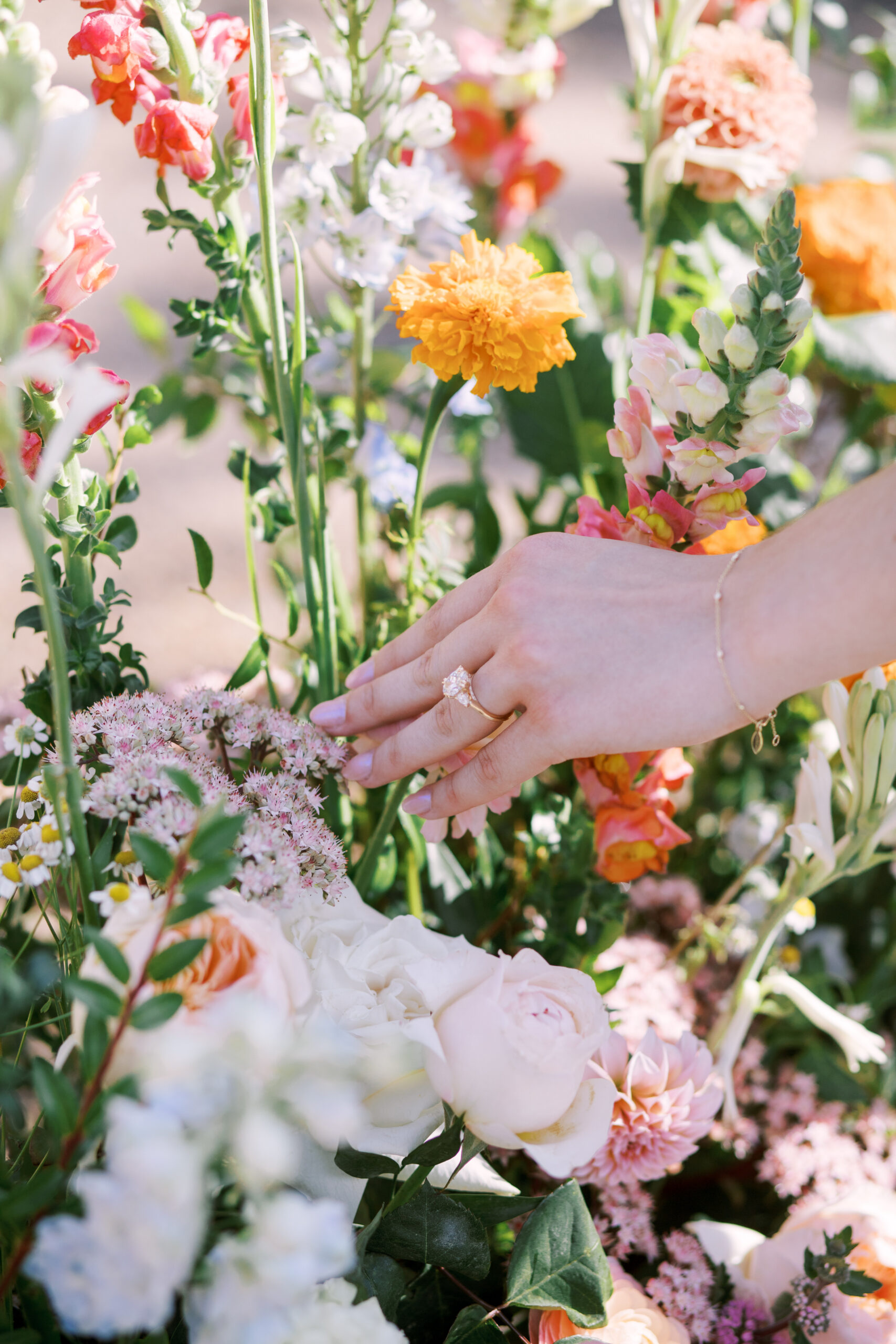 Close-up of engagement ring on woman’s hand among soft pastel garden flowers.