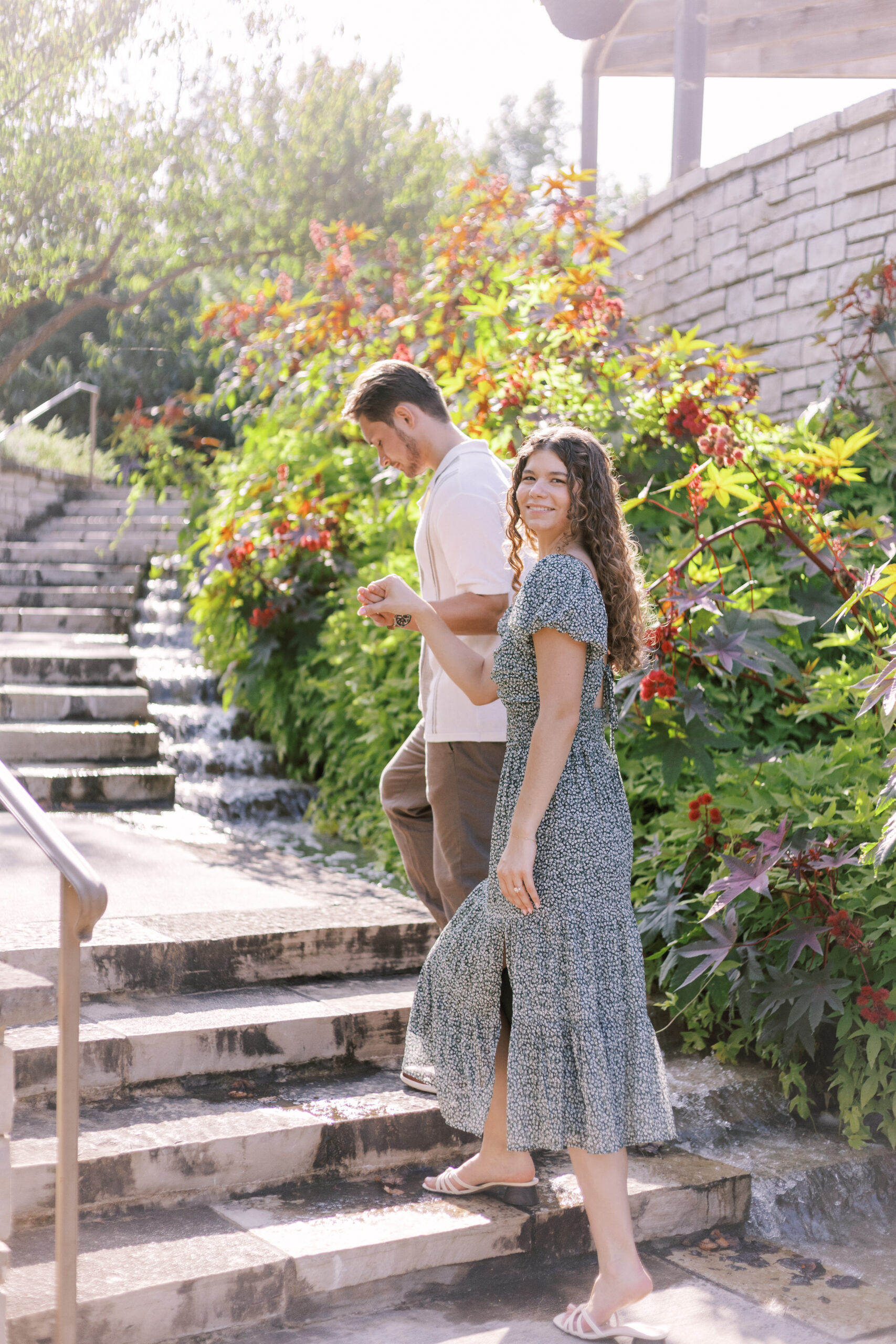 Couple walking up sunlit garden steps, woman glancing back, after engagement at Powell Gardens.