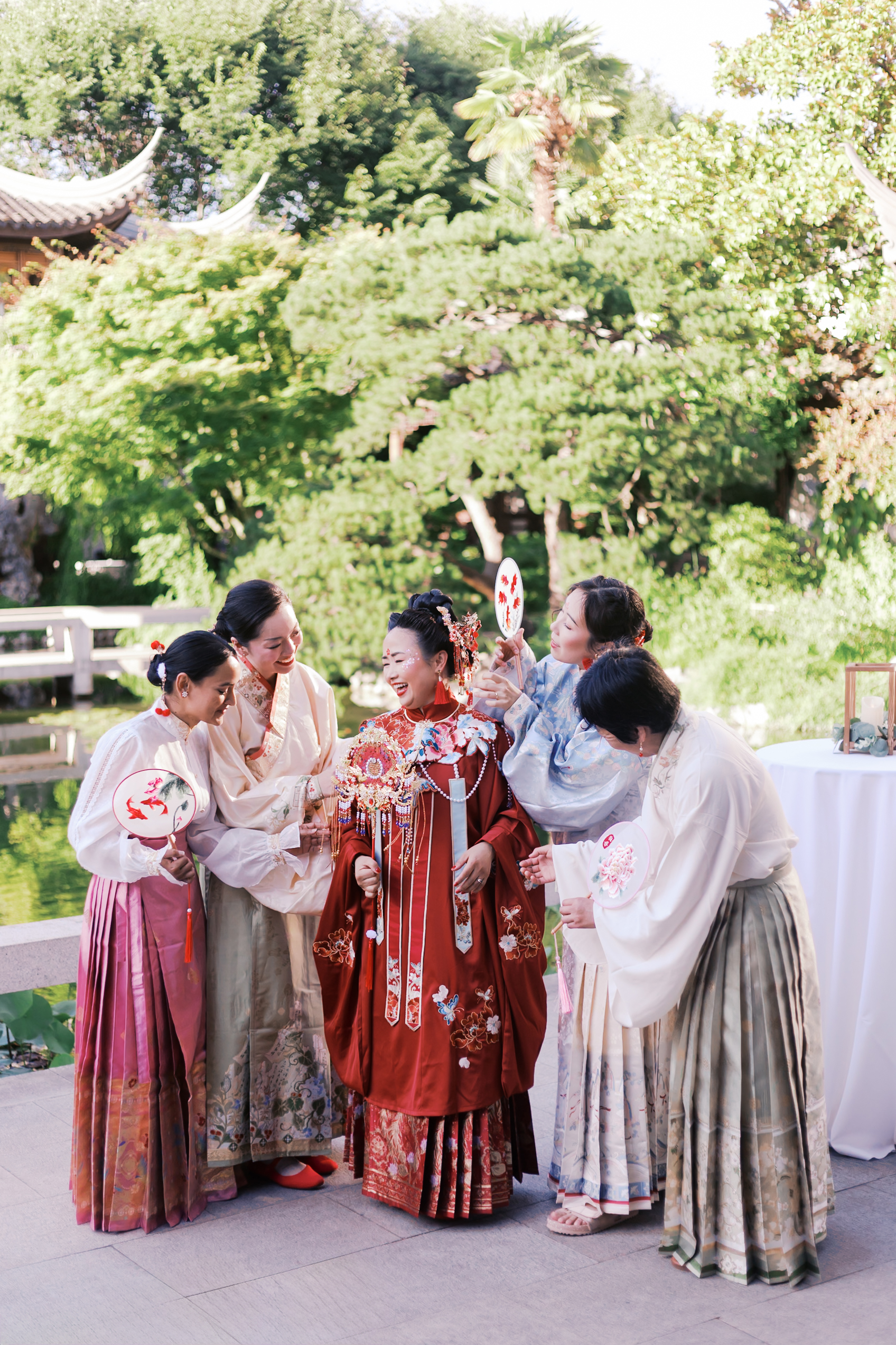 Bride in red traditional Chinese wedding dress surrounded by bridesmaids in pastel hanfu dresses at a garden venue.