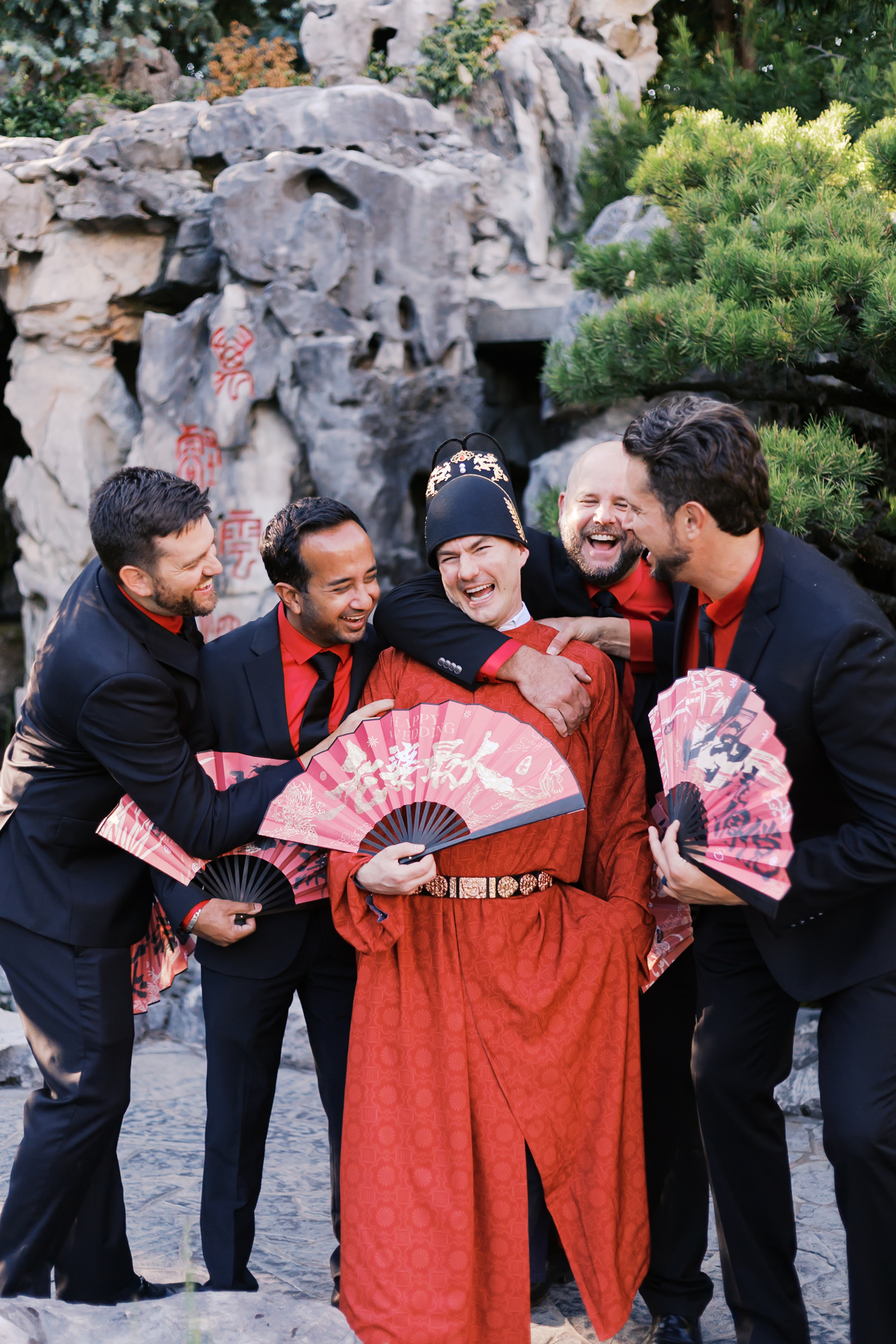 Groom in traditional red Chinese wedding attire laughing with groomsmen holding red folding fans in a garden courtyard.