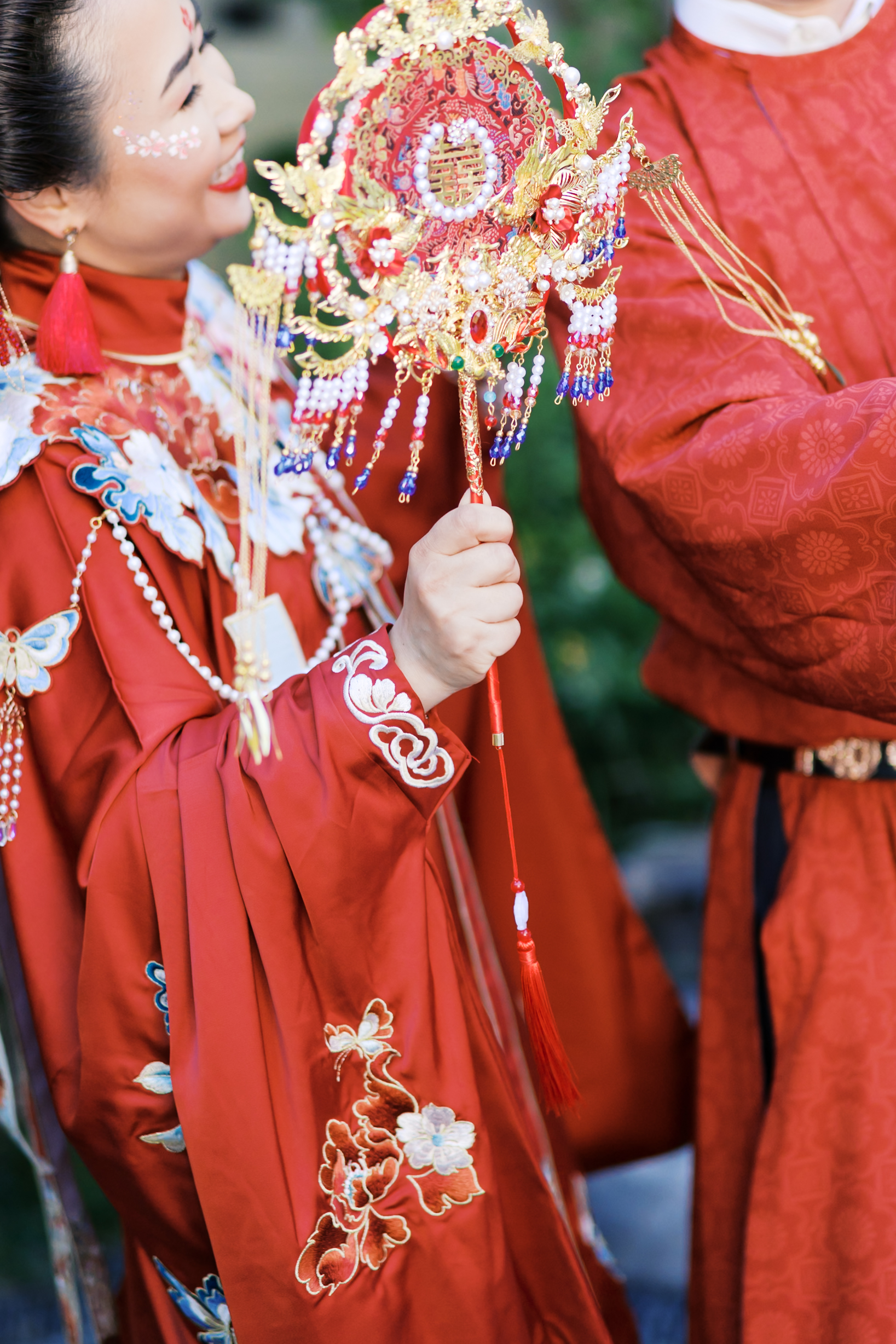 Bride holding an ornate red and gold Chinese bridal fan while wearing embroidered traditional wedding robes beside groom.