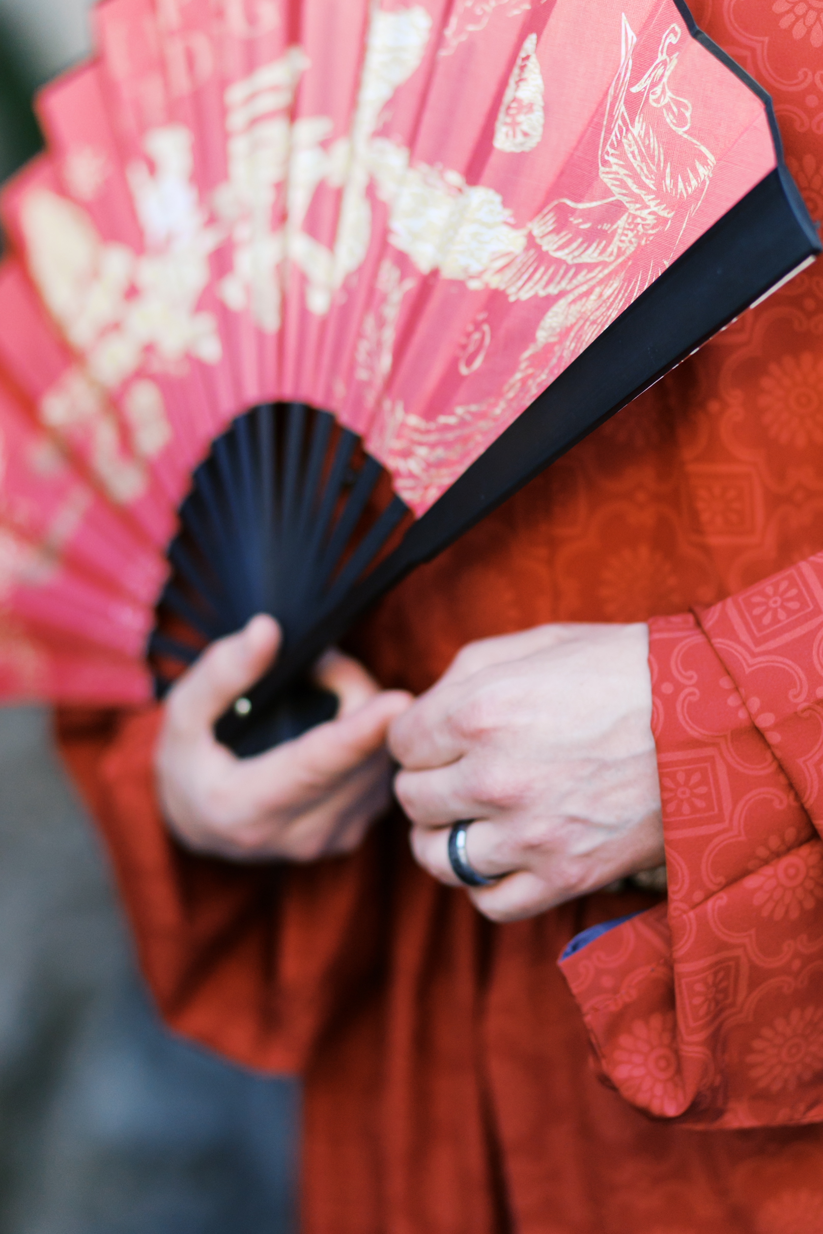 Close-up of groom’s hands holding a red folding fan with gold Chinese wedding artwork and traditional red robes.
