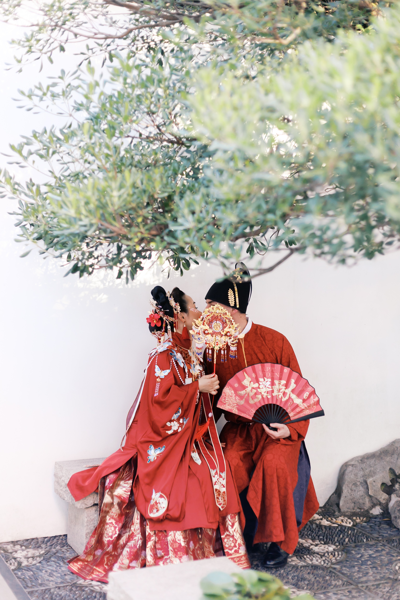 Couple in traditional Chinese wedding attire sharing a kiss beneath green trees while holding decorative wedding fans.