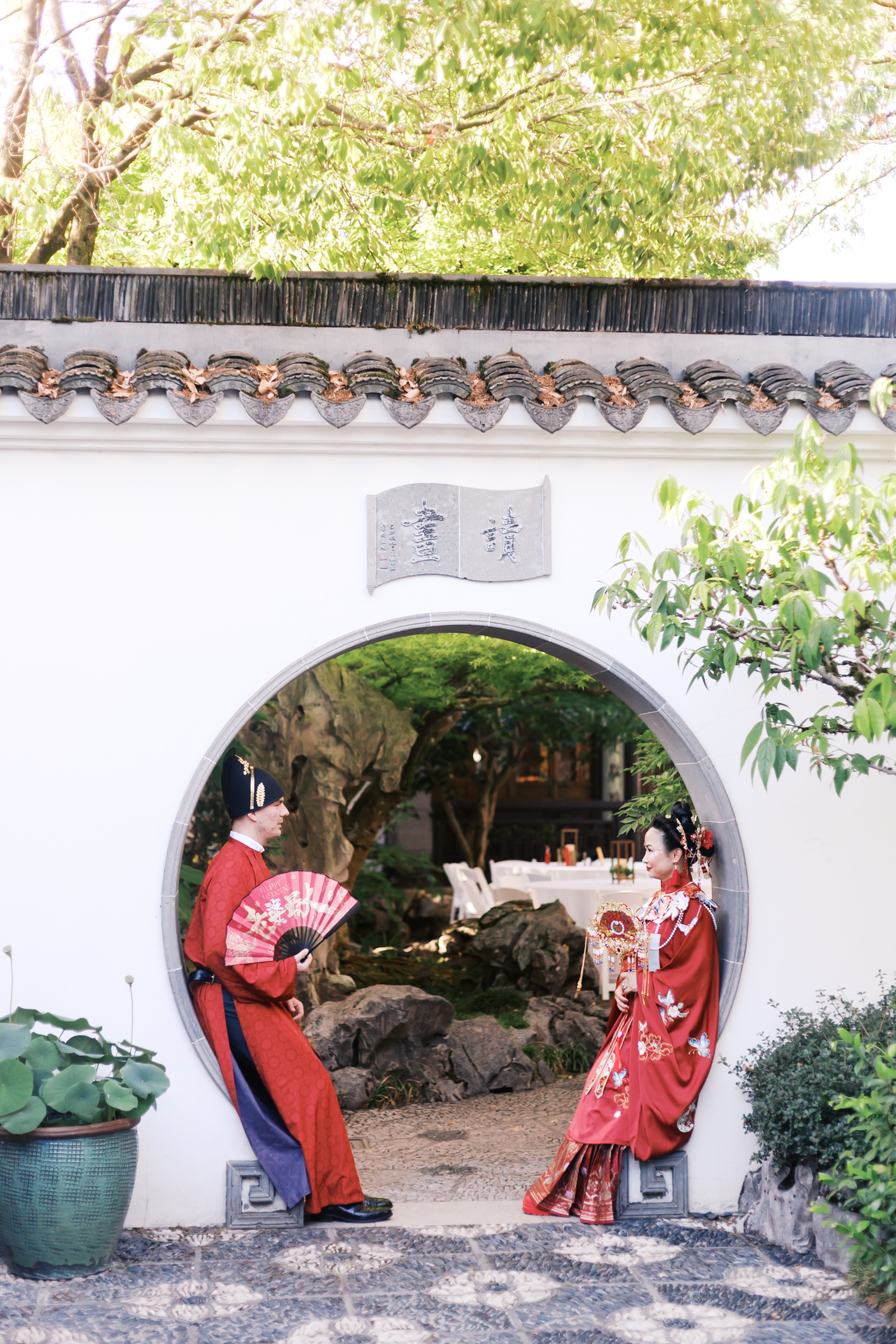 Bride and groom in red traditional Chinese wedding clothing standing beneath a circular moon gate in a garden courtyard.