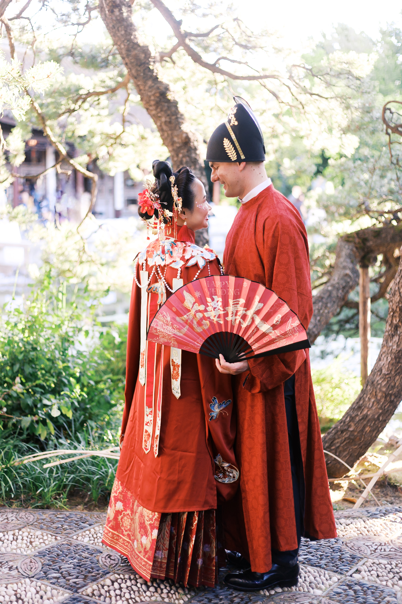 Couple in embroidered red Chinese wedding attire posing together in a sunlit garden with decorative folding fan.