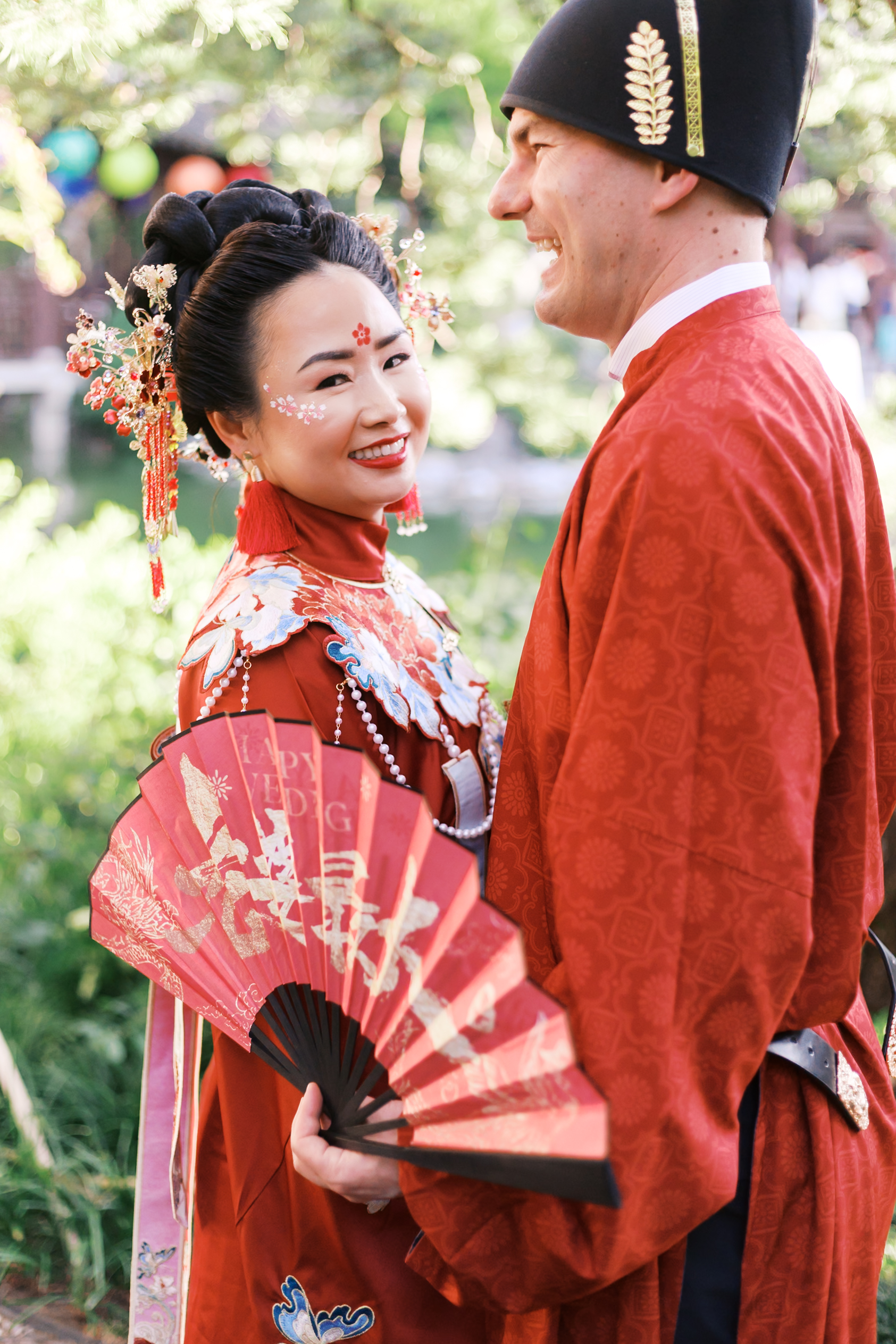 Bride smiling toward camera beside groom while holding a red Chinese wedding fan in a garden setting.