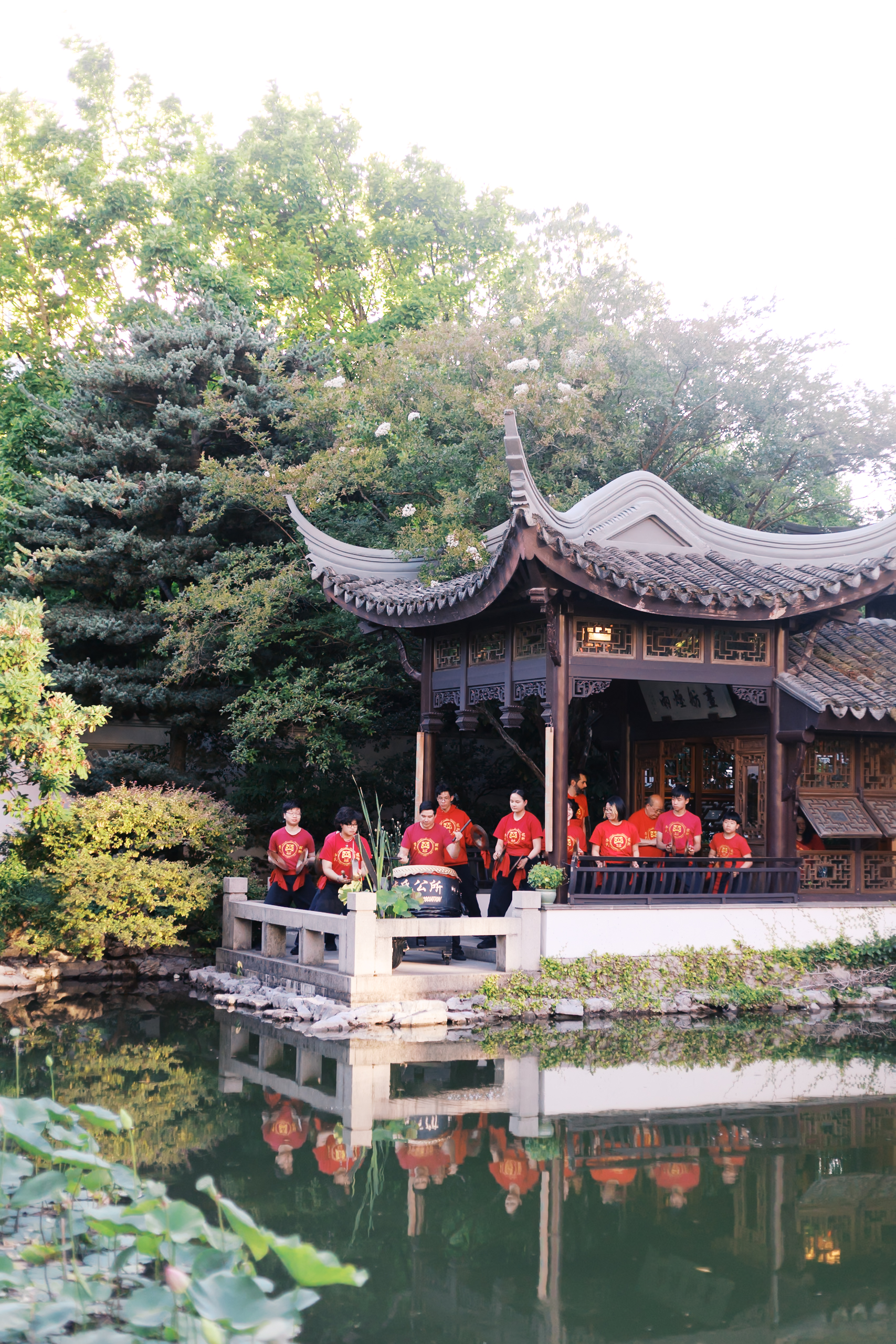 Traditional Chinese drum performers in red shirts performing beside a pavilion overlooking a garden pond.