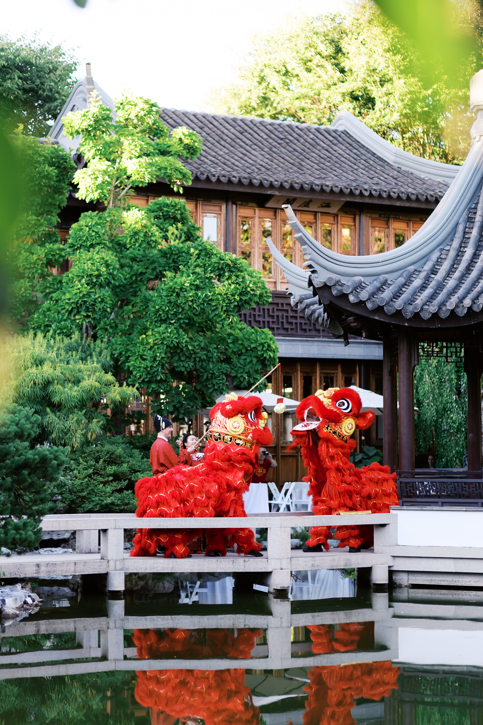 Two red lion dance performers entertaining wedding guests beside a pond and traditional Chinese architecture.