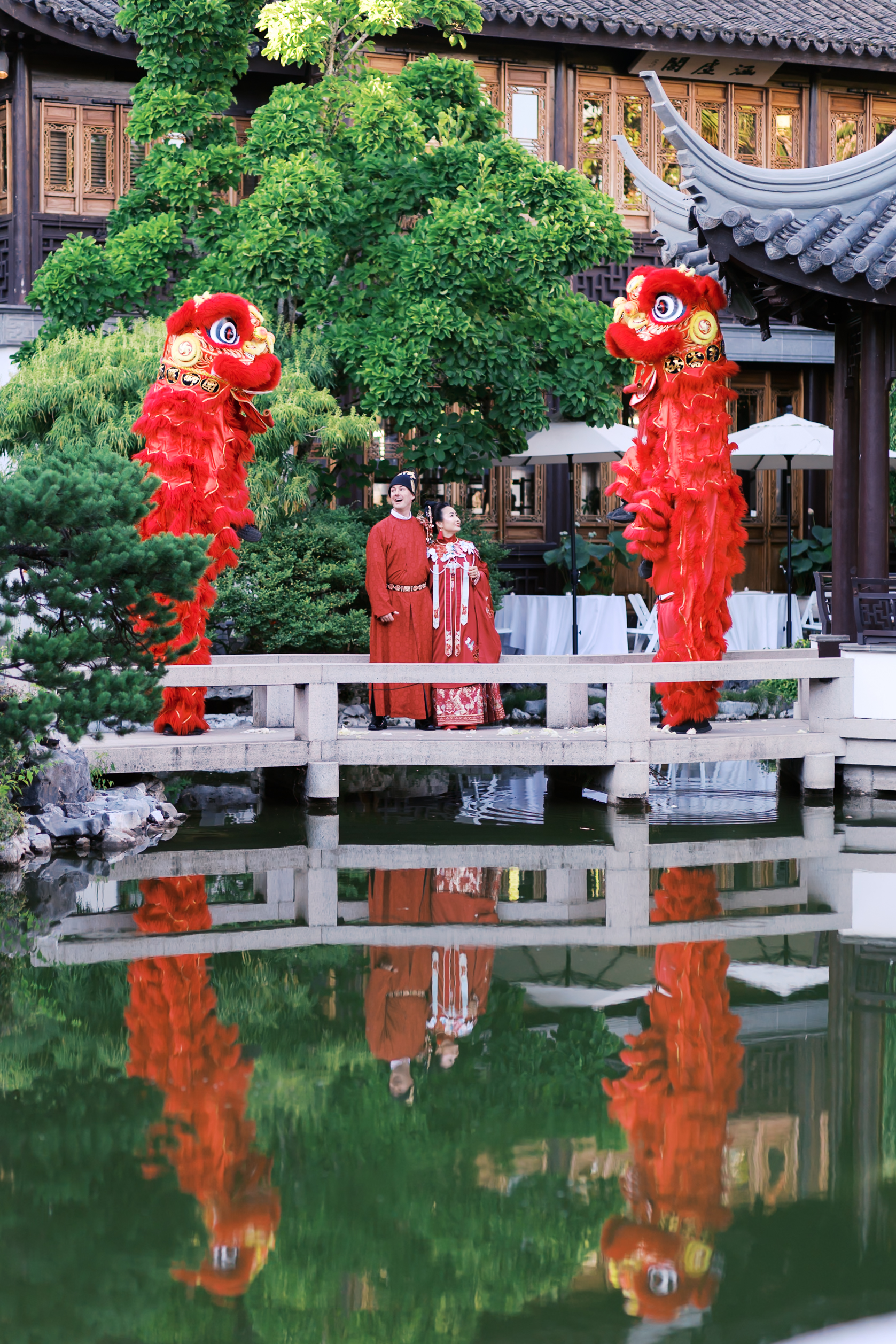 Bride and groom watching lion dancers perform on a bridge in a Chinese garden wedding venue.