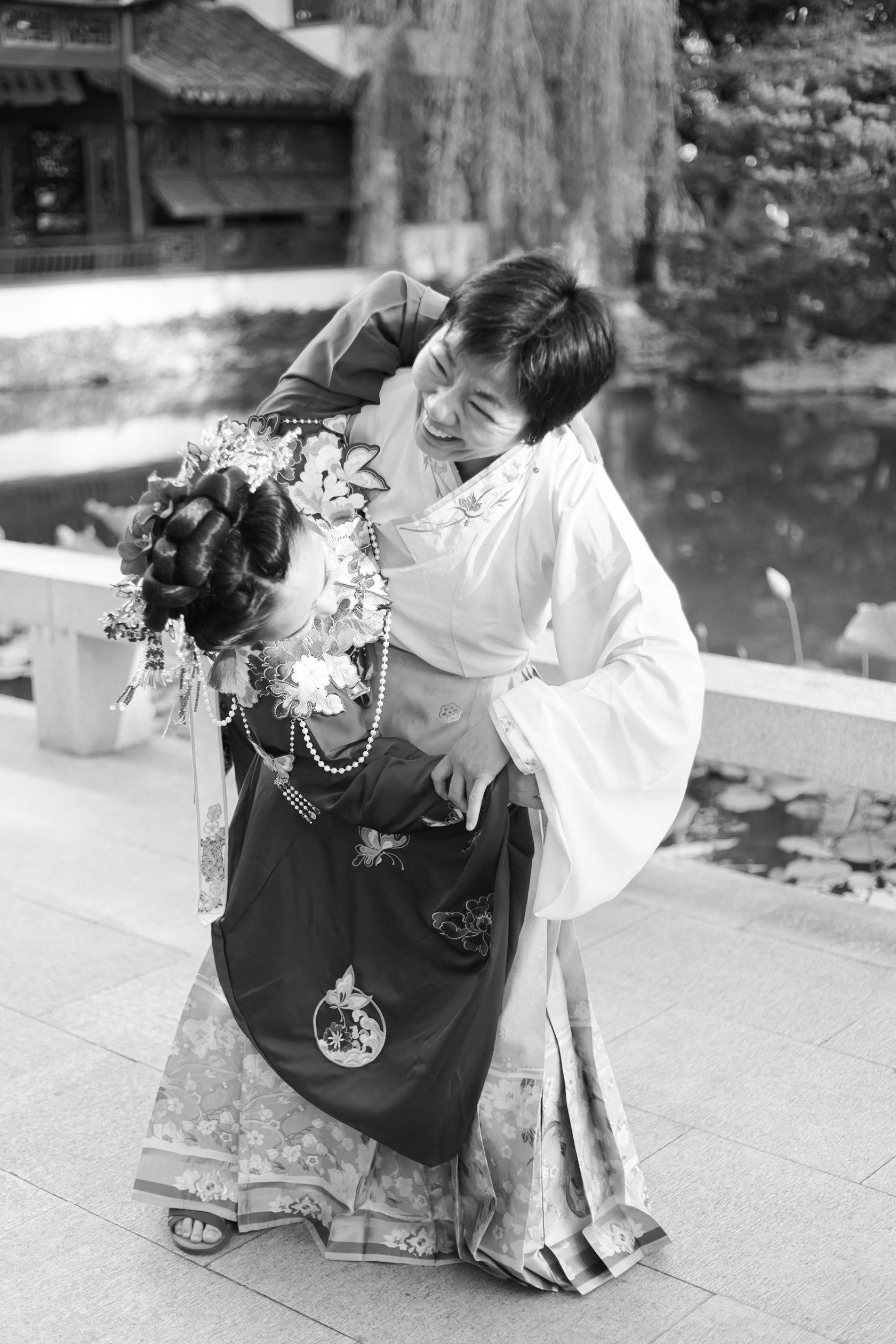 Black-and-white portrait of woman dipping bride in a traditional Chinese wedding outfit beside a pond.
