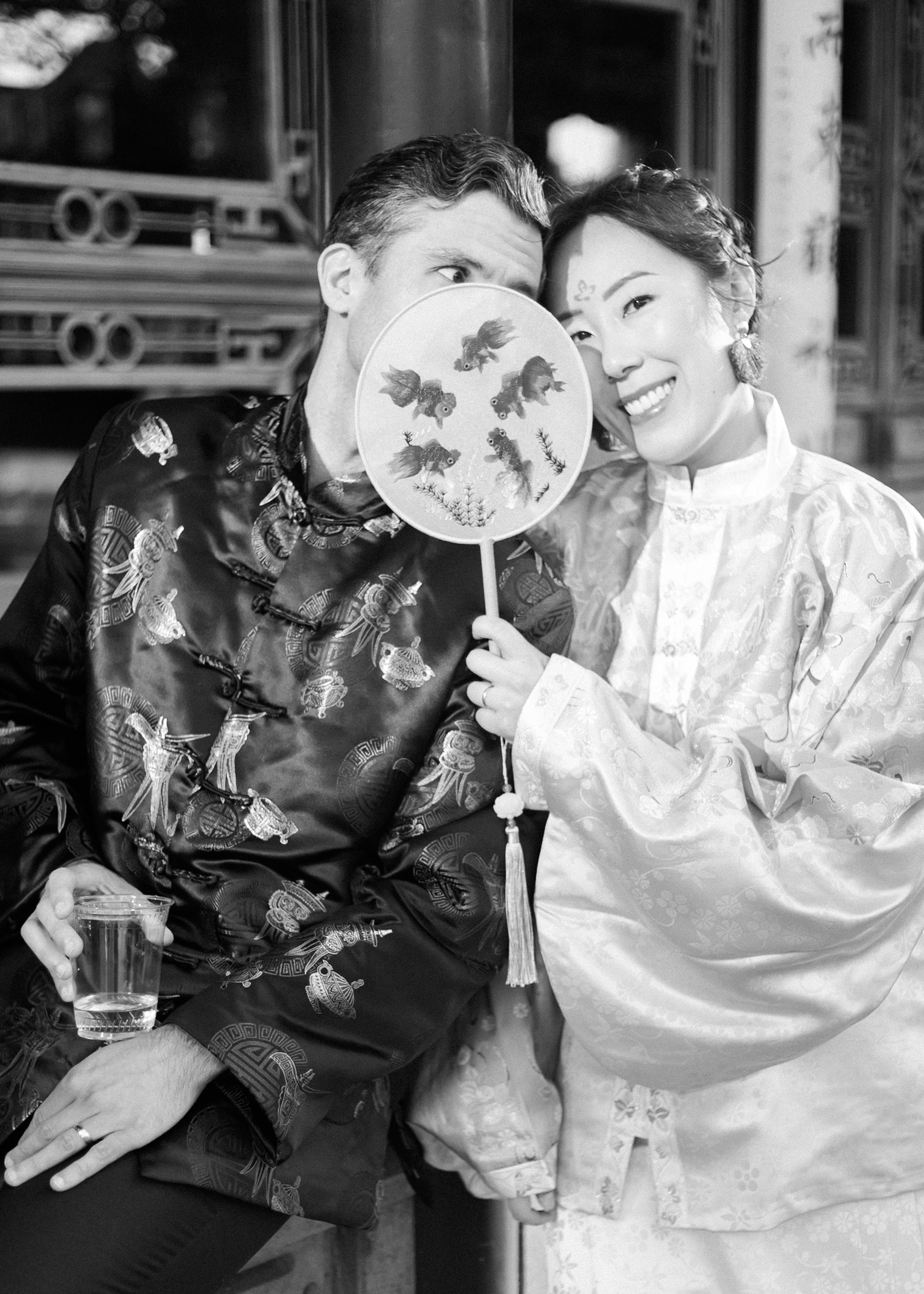 Black-and-white portrait of couple in traditional Chinese clothing posing with decorative embroidered fan.
