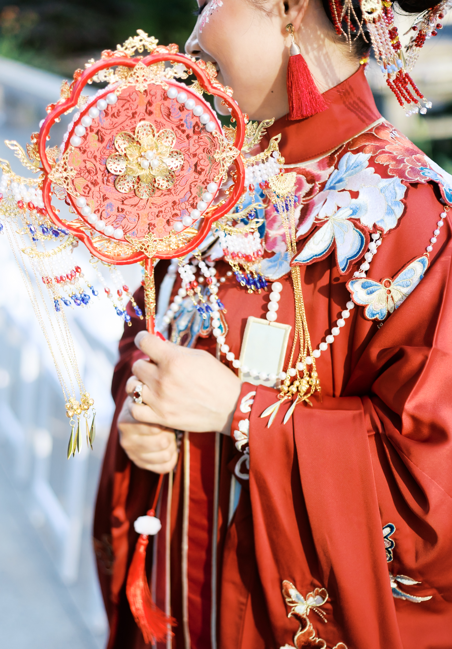 Close-up of bride in ornate red Chinese wedding dress holding embroidered circular fan with tassels.