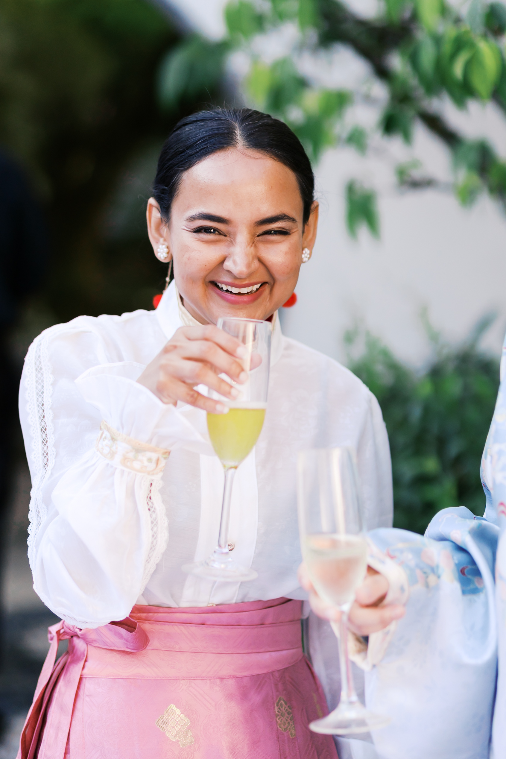 Smiling wedding guest in white and pink hanfu raises a champagne glass at an outdoor garden celebration.