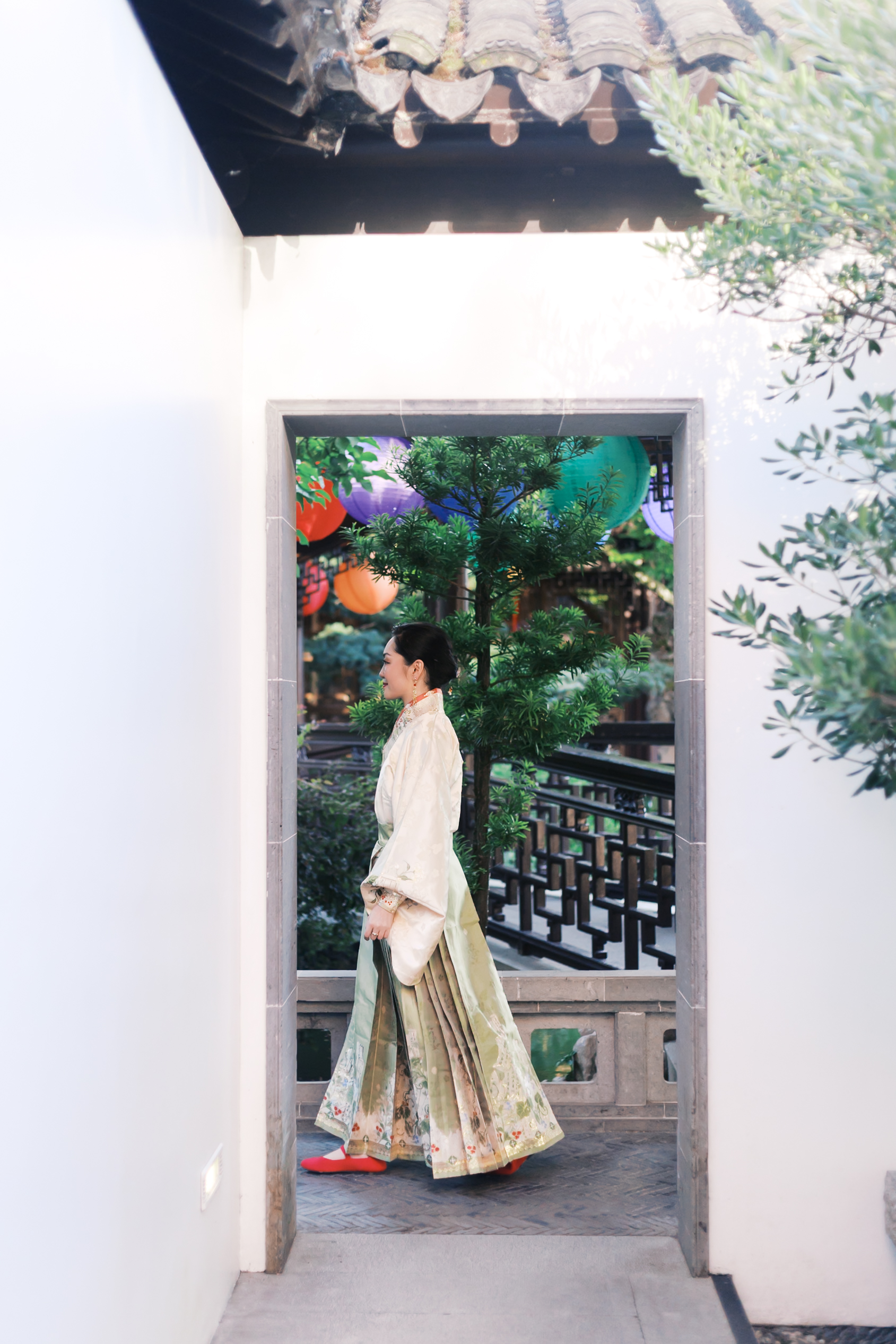Woman in embroidered hanfu walking through a moon gate framed by lanterns and garden architecture.