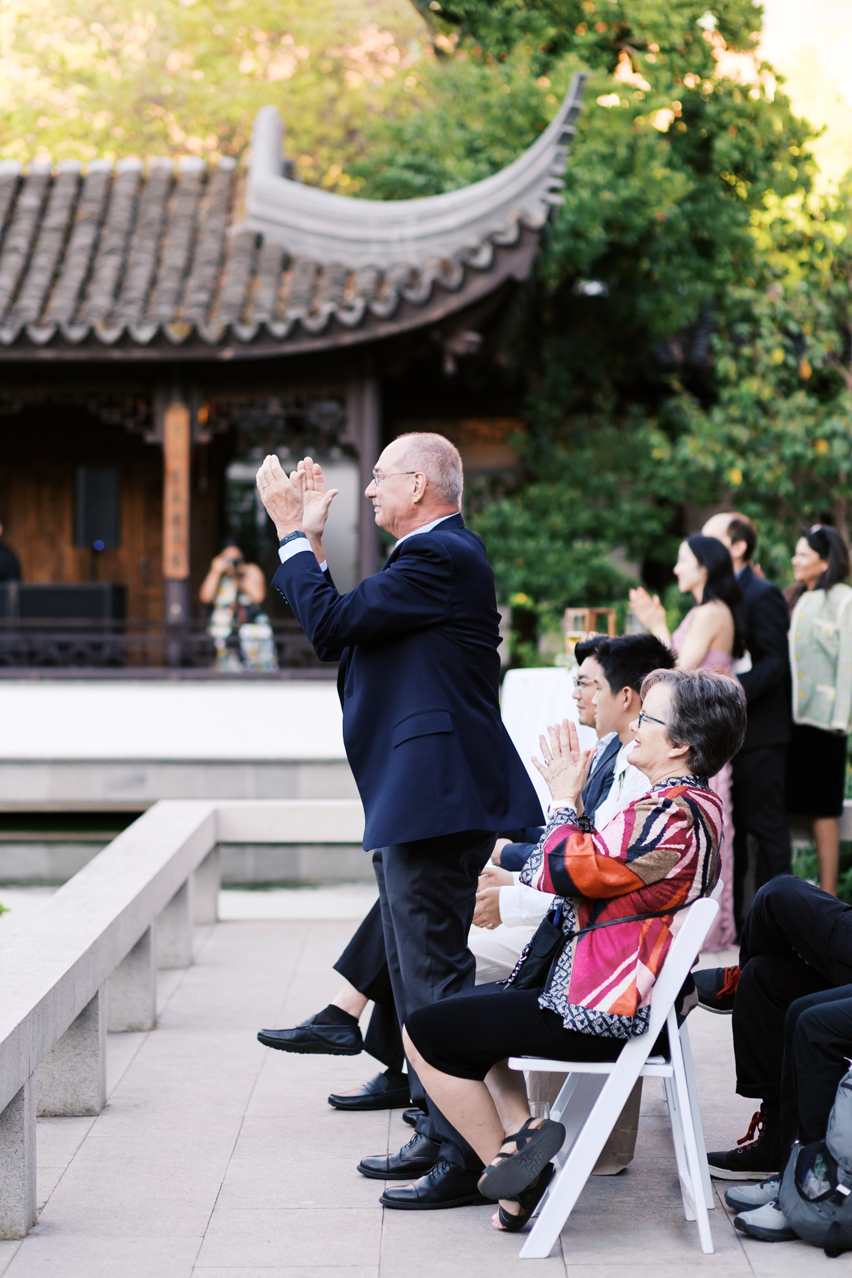 Wedding guests applauding during an outdoor reception beside a traditional Chinese pavilion.