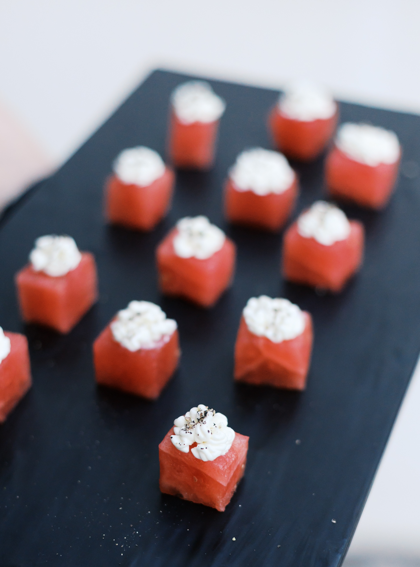 Bite-sized watermelon appetizers topped with cream cheese and seeds on a black serving tray.