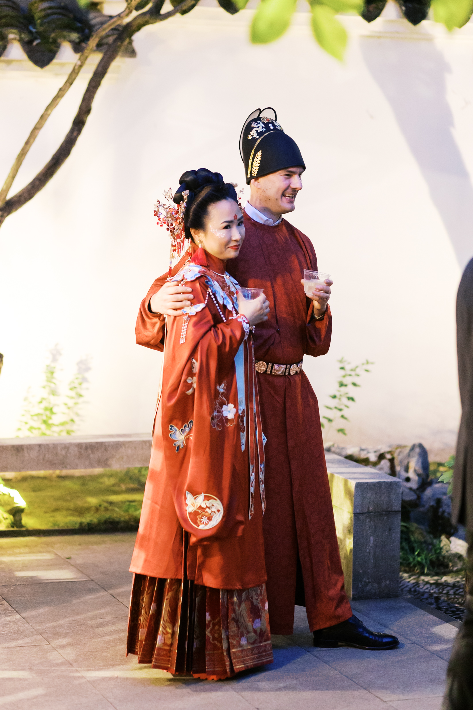 Couple in matching red traditional Chinese wedding outfits holding drinks during evening reception.