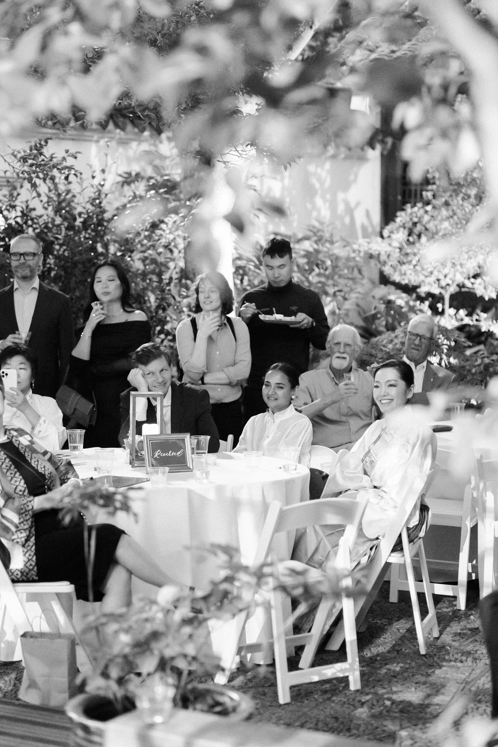 Black-and-white photo of wedding guests seated outdoors listening to speeches at a garden reception.
