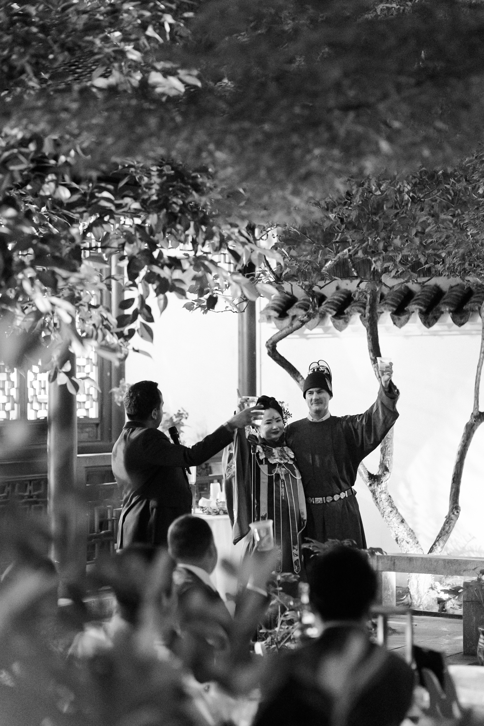 Black-and-white image of bride and groom raising glasses for a toast during wedding reception speeches.